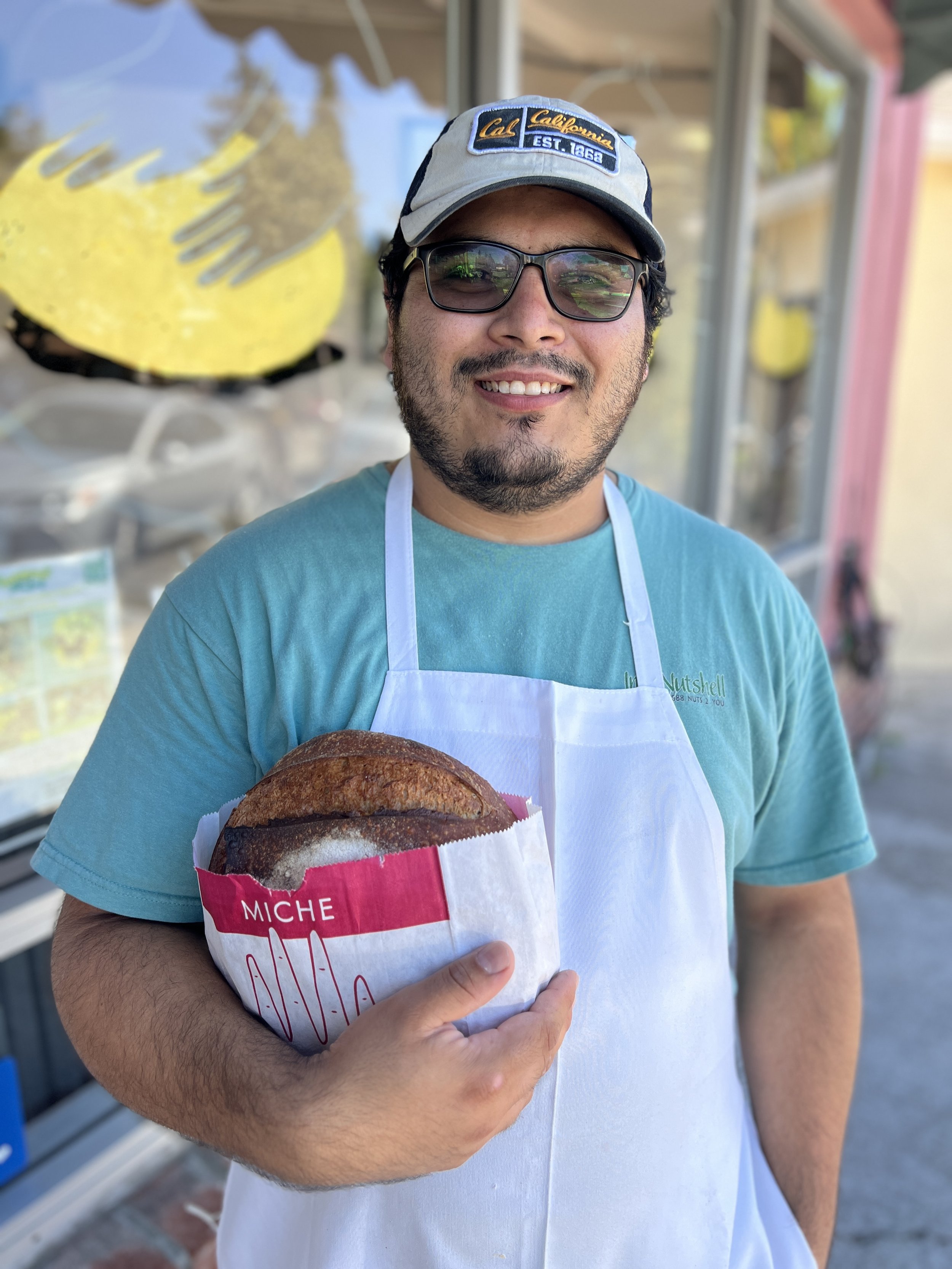 A smiling man wearing glasses, a cap with "Cal California EST. 1868" patch, and a white apron is holding a large sandwich wrapped in white and pink paper, outside a storefront with colorful decorations.