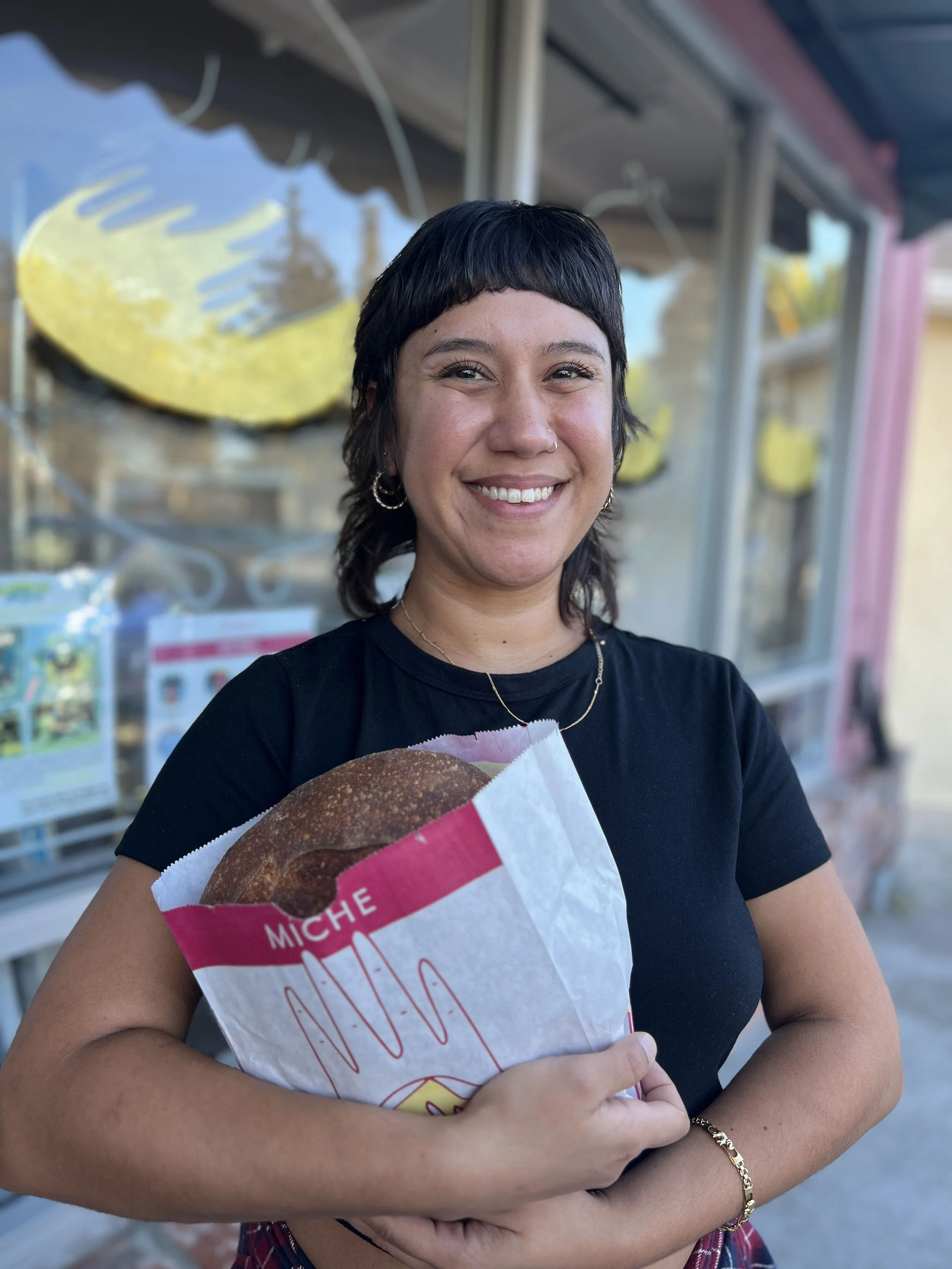 A woman smiling and holding a sandwich wrapped in white and pink paper outside a store or stand.