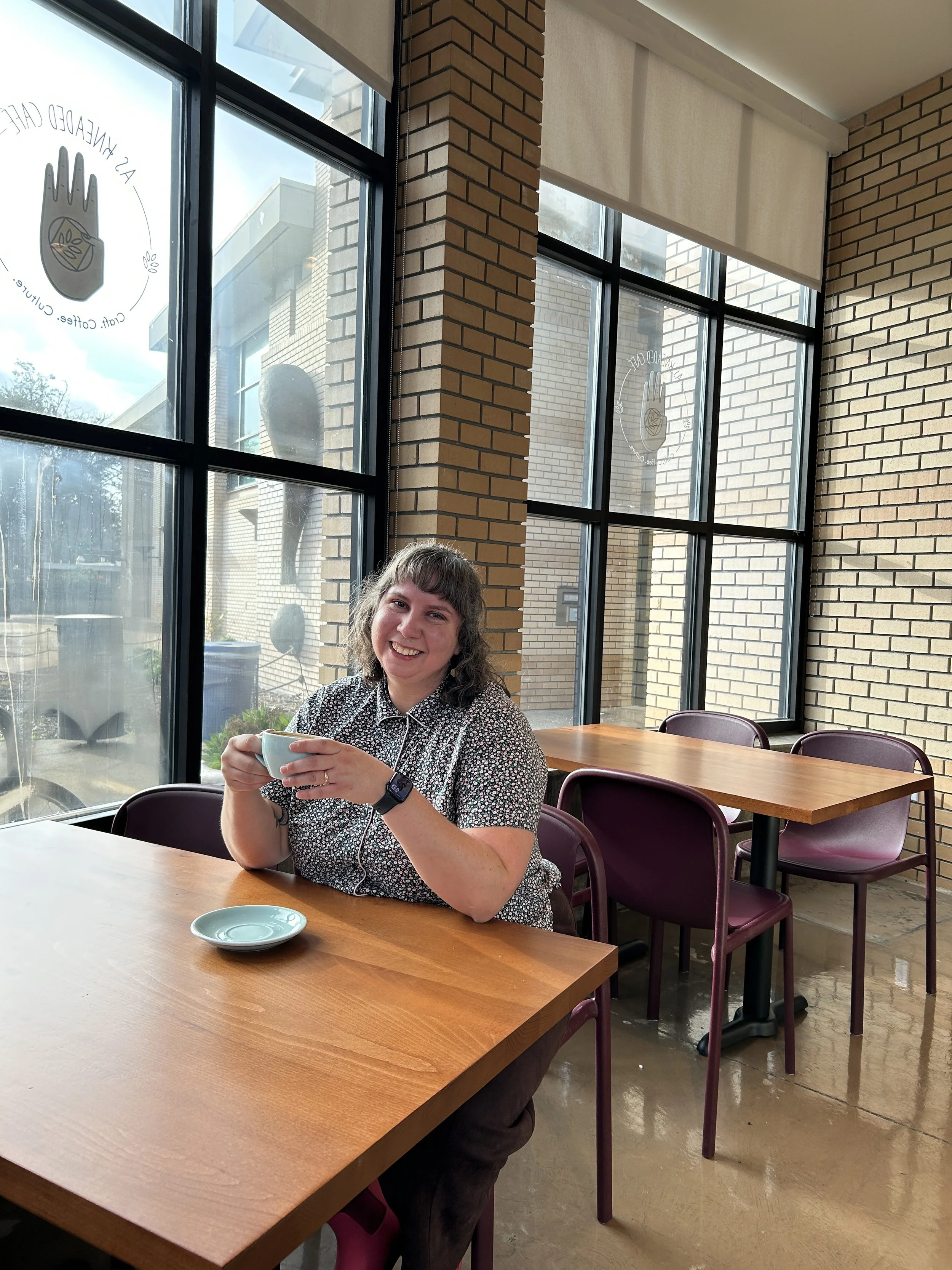 A smiling woman with short curly hair, wearing a patterned shirt, sitting at a wooden table in a cafe, holding a teal mug. There are empty teal saucers on the table, large windows with a brick wall exterior, and purple chairs.