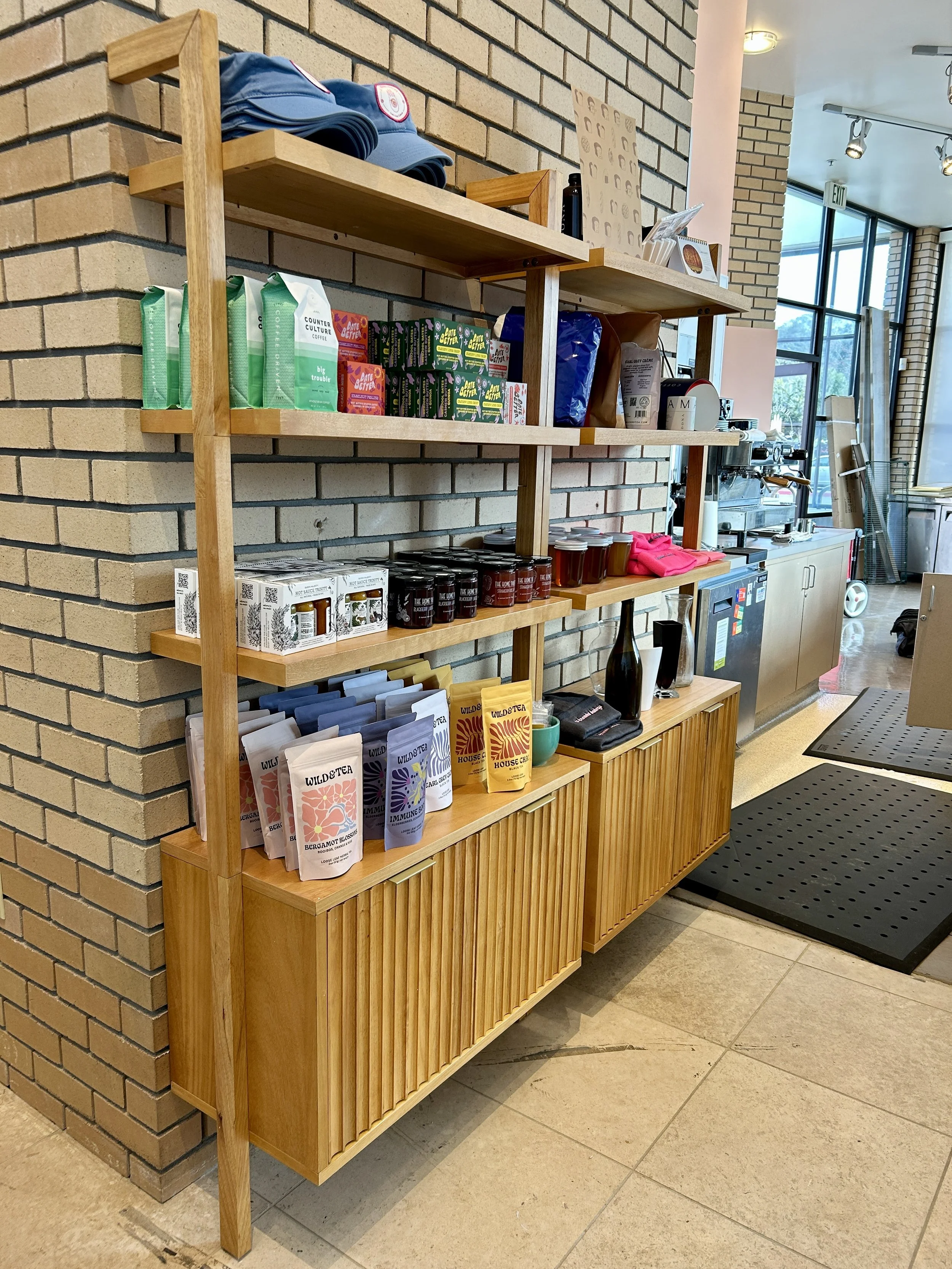 Wooden shelving unit with coffee and tea supplies, including tea bags, coffee, mugs, and jars, located in a café near a brick wall.