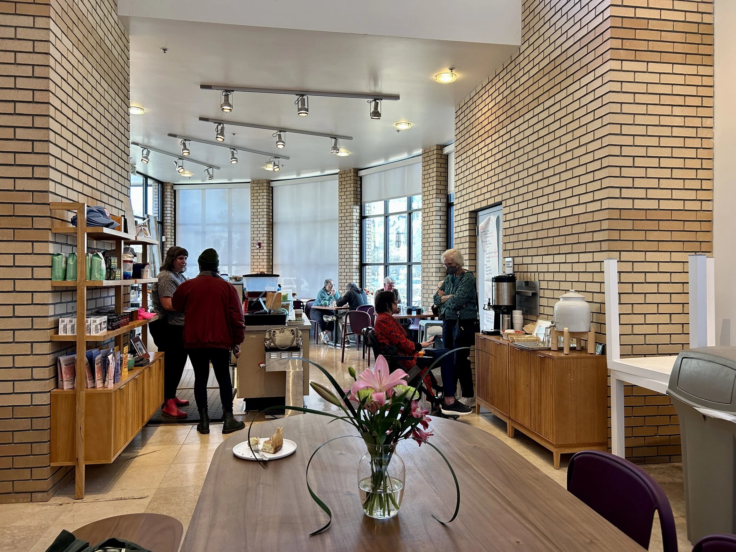 Inside a coffee shop or cafe with brick walls, large windows, and several people seated and standing, some wearing masks. There is a wooden table with a vase of pink flowers in the foreground and a breakfast plate with a pastry and fruit on a table.