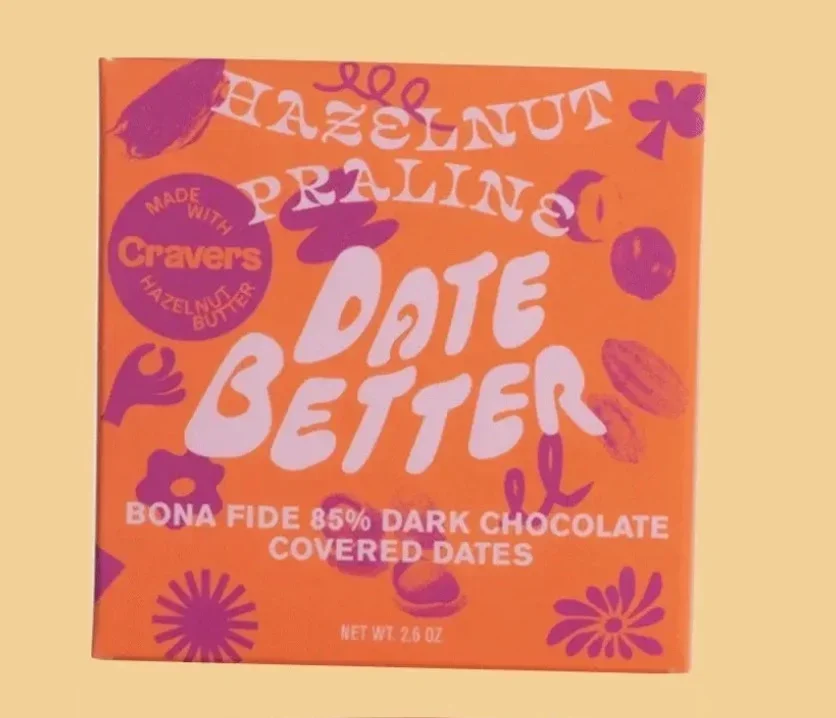 Orange packaging of Date Better chocolate with purple and white text, featuring colorful illustrations of dates, flowers, and a logo indicating it's made with Cravers hazelnut butter. The box states it contains 85% dark chocolate-covered dates, net weight 2.6 ounces.