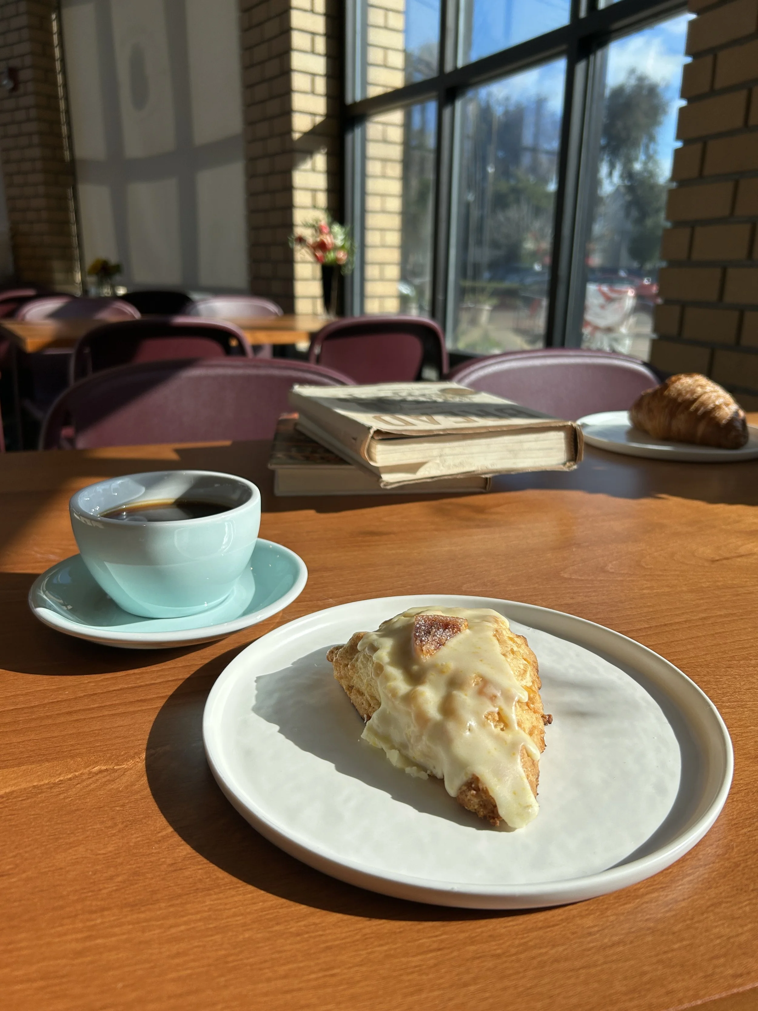 A scone with icing on a white plate, a cup of black coffee in a light blue cup and saucer, a croissant on a white plate, and a book on a wooden table in a sunlit café with pink chairs and a large window.