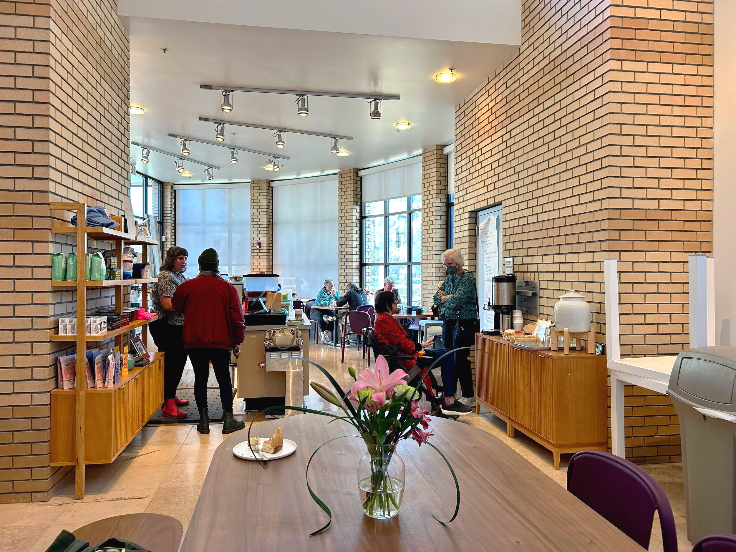 Interior of a cafe or restaurant with brick walls, large windows, and a small counter area. Several people are sitting at tables, and some are standing and talking. There is a vase with pink flowers on the foreground table.