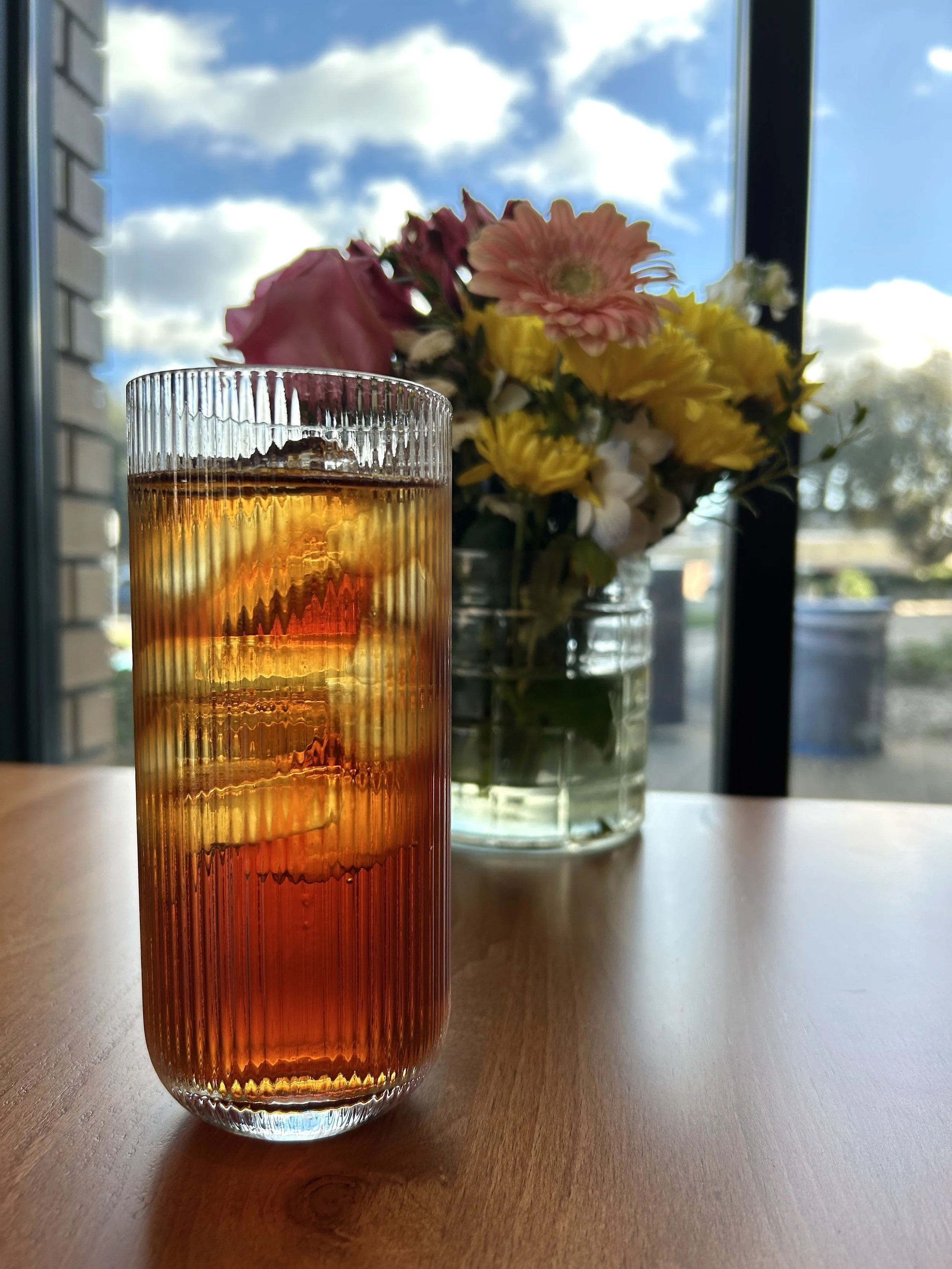 Glass of iced tea with lemon slices on a wooden table, with a bouquet of multicolored flowers in the background beside a window showing a partly cloudy sky.