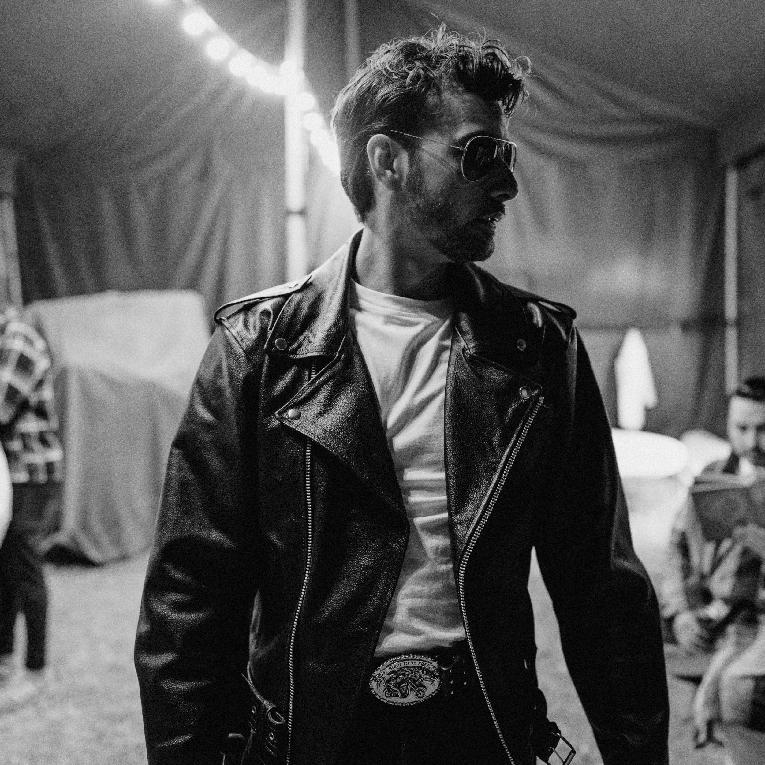 A man with a beard and sunglasses wearing a leather jacket and white t-shirt, looking to the side, in an indoor setting with soft lighting and people in the background.