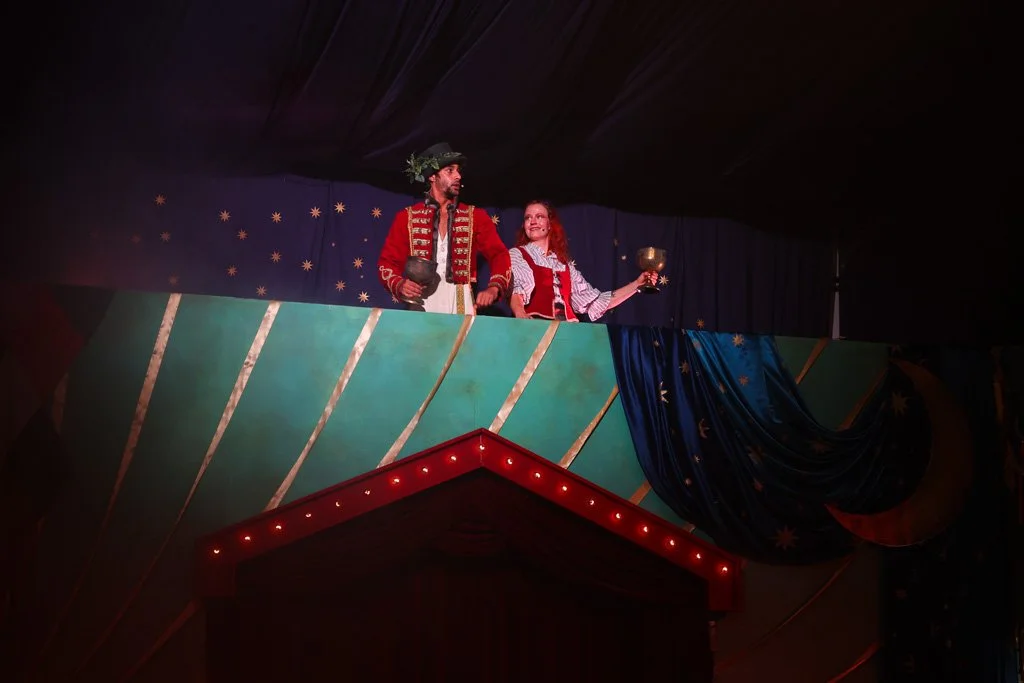 Two performers in colorful theatrical costumes stand on a stage balcony, with one holding a goblet and the other a cup, under a dark ceiling with decorated drapes and star patterns.