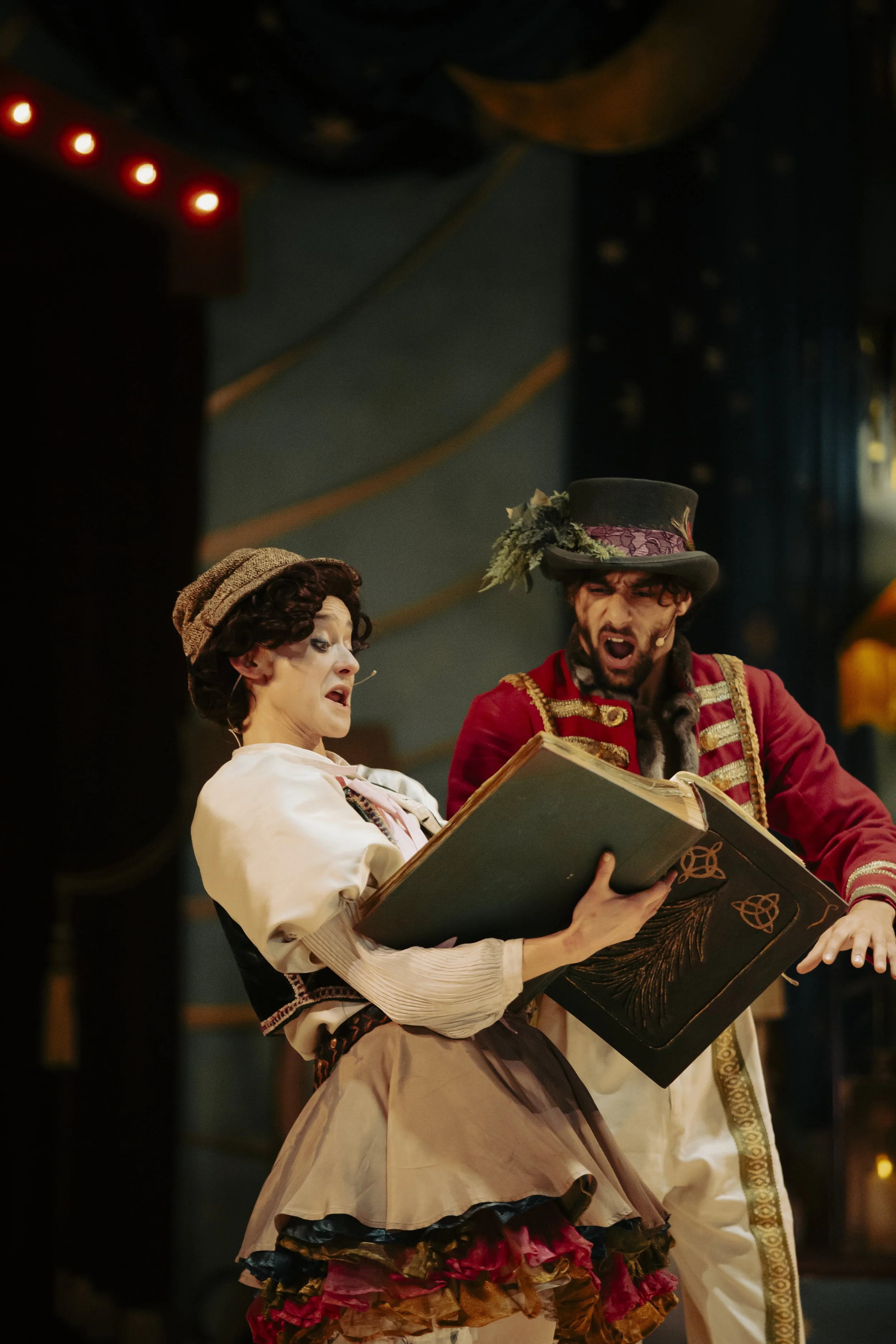 Two actors performing on stage, dressed in elaborate costumes; one with a brown wig and hat, holding a large book, and the other with a black hat decorated with flowers, singing passionately.