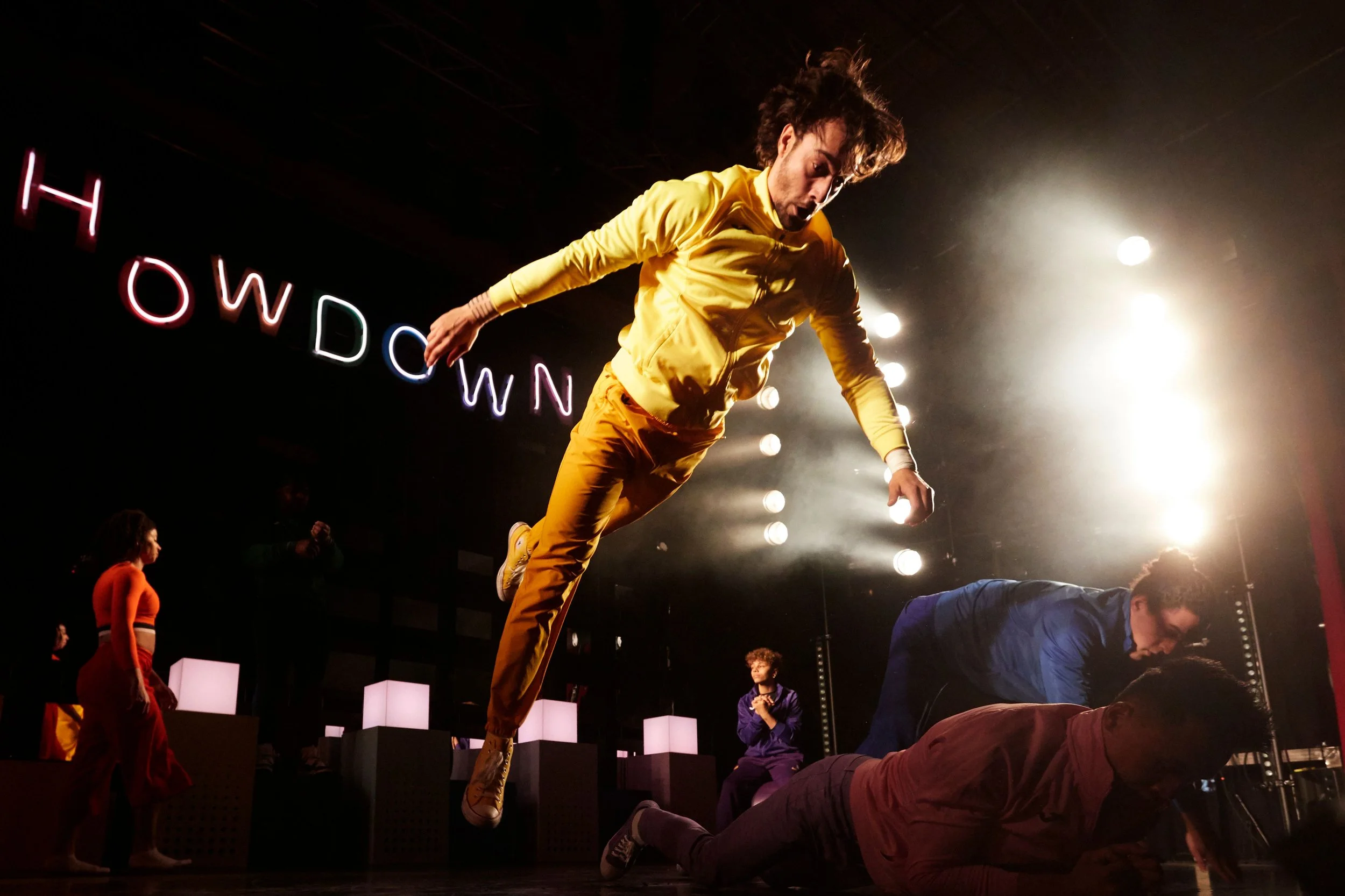 A performer in a yellow outfit jumps in the air during a stage performance, with other performers and stage lights in the background.