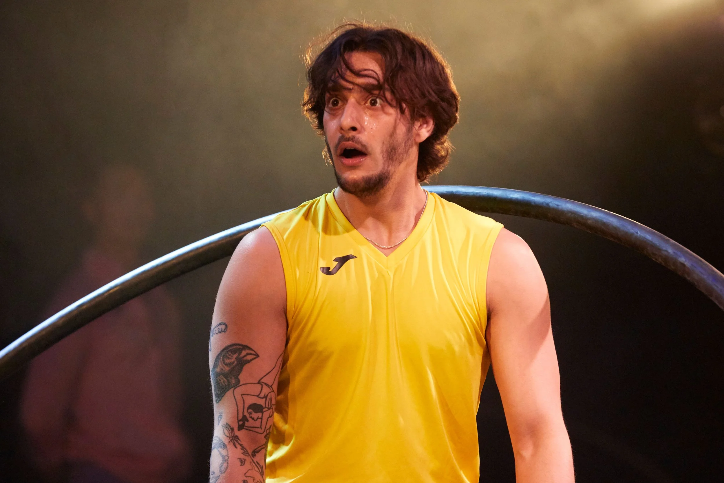 A man with curly brown hair and a beard, wearing a yellow sleeveless athletic shirt, looks surprised or shocked, with a dark background and a curved metal bar behind him.