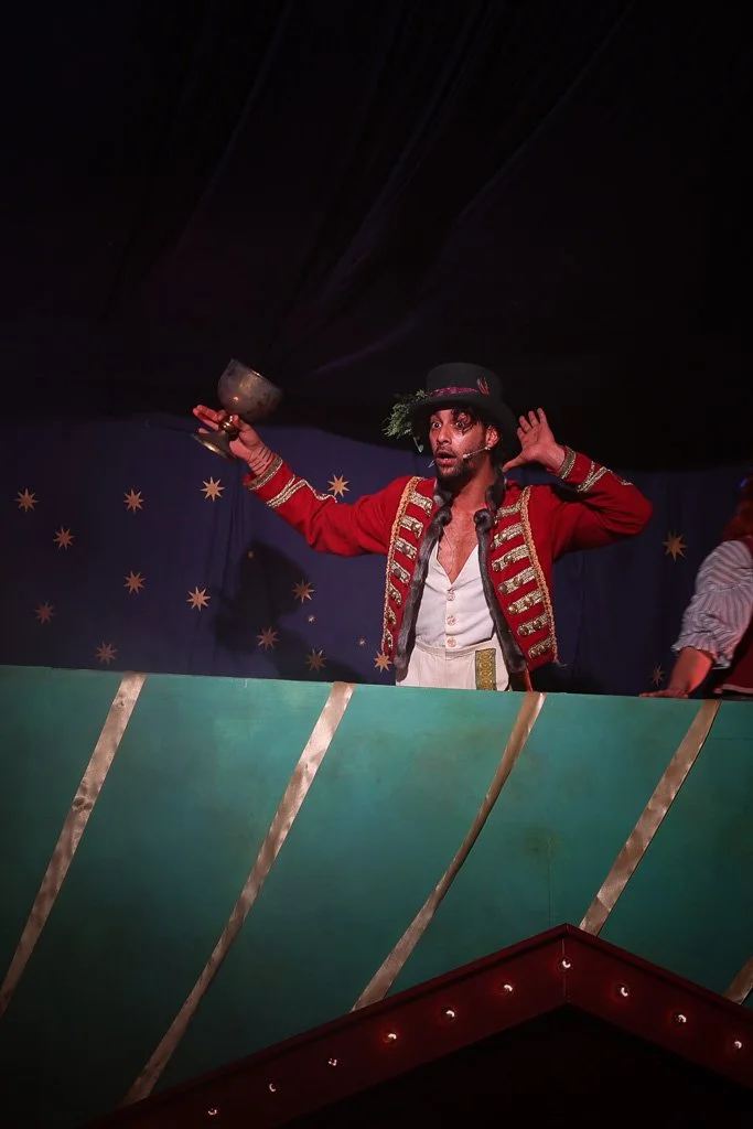 Performer dressed as a circus ringmaster in a red coat with gold trim, black hat, holding a small bowl or cup, with a dark draped backdrop decorated with gold stars.