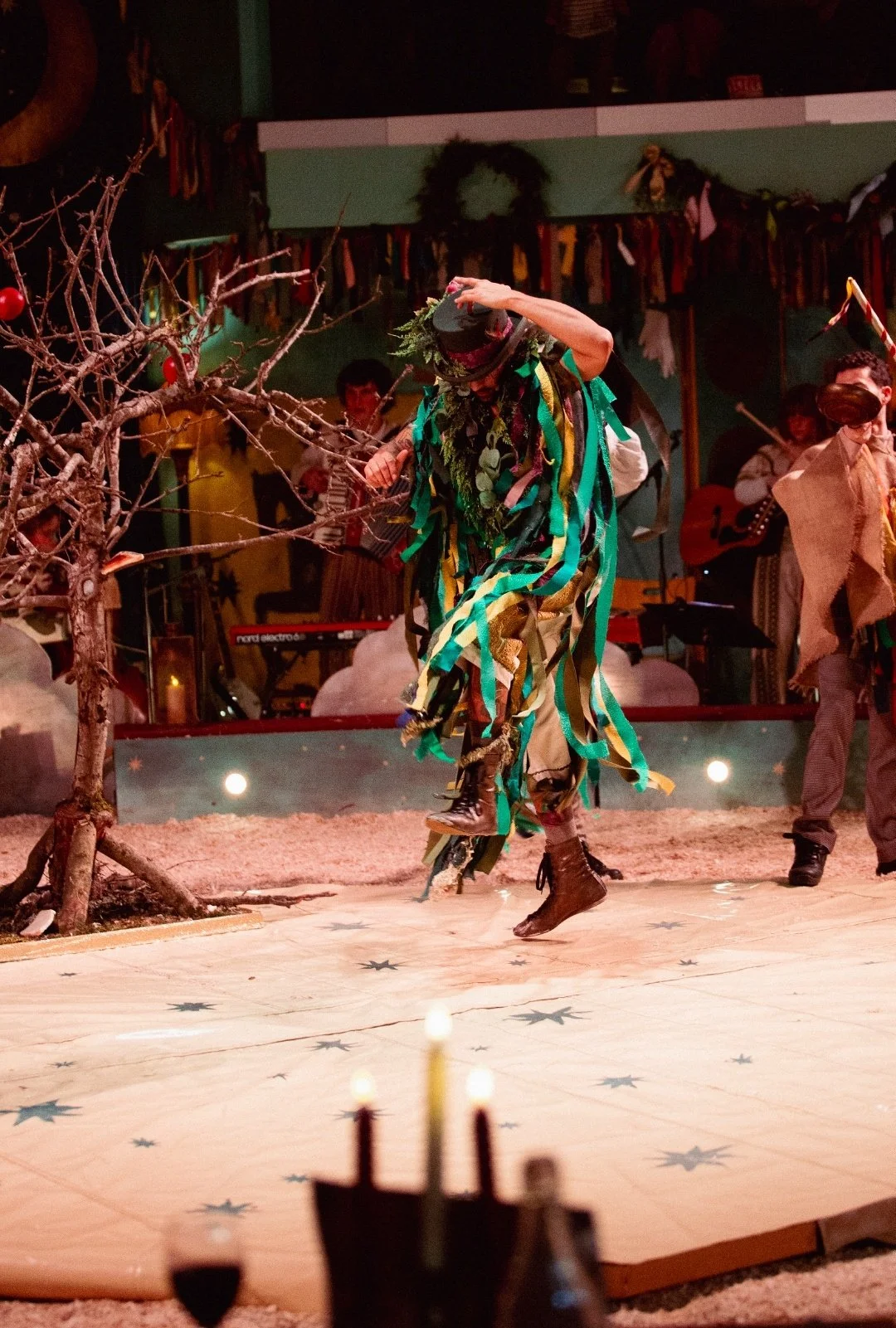 Performer dancing in a colorful costume made of ribbons with a black hat decorated with greenery, on a stage decorated with a leafless tree and stars, with musicians playing in the background.