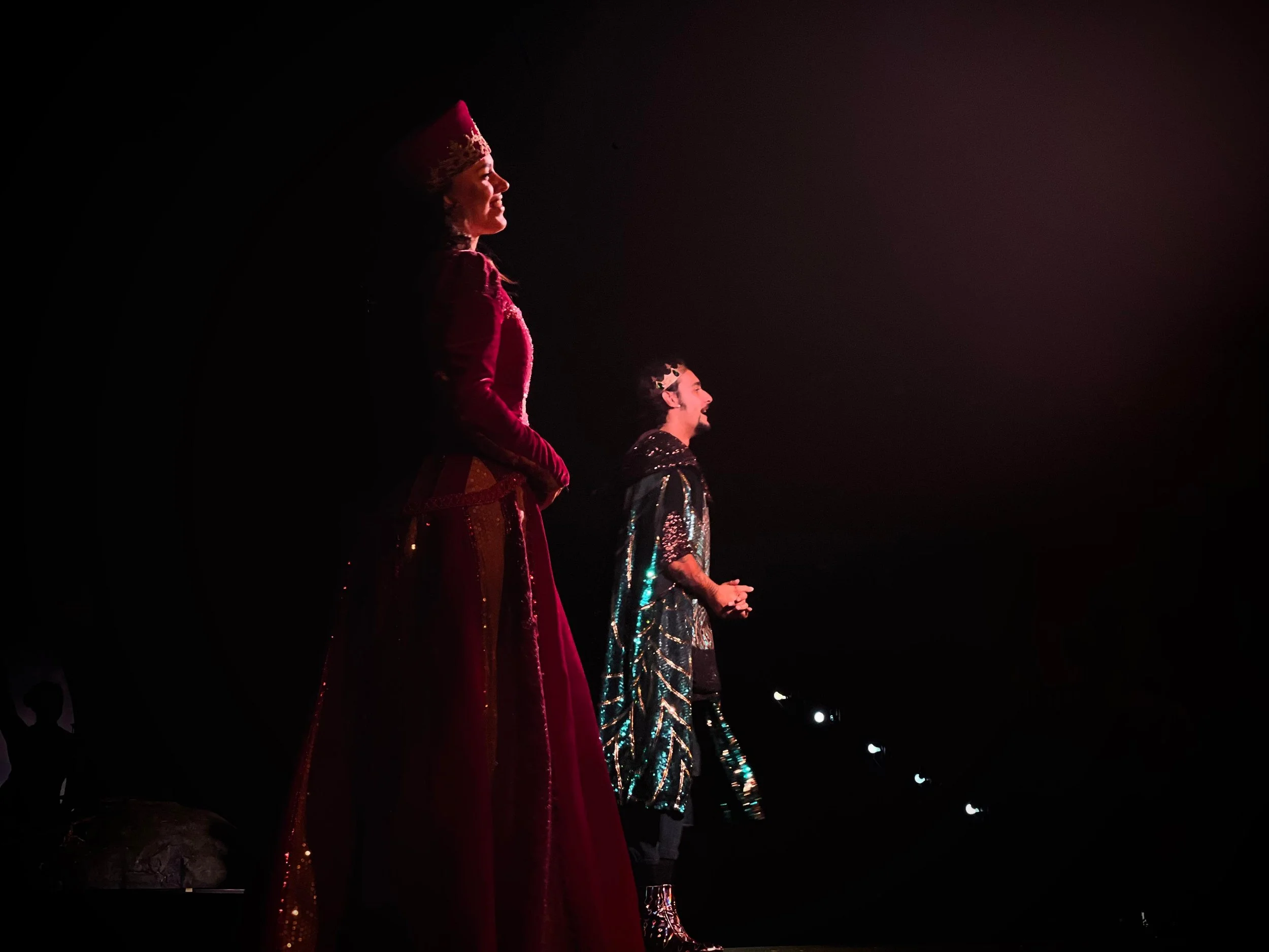 Two performers dressed in elaborate costumes stand on stage in profile, illuminated by stage lights against a dark background, with a woman in a red gown and a man in a black and gold outfit.