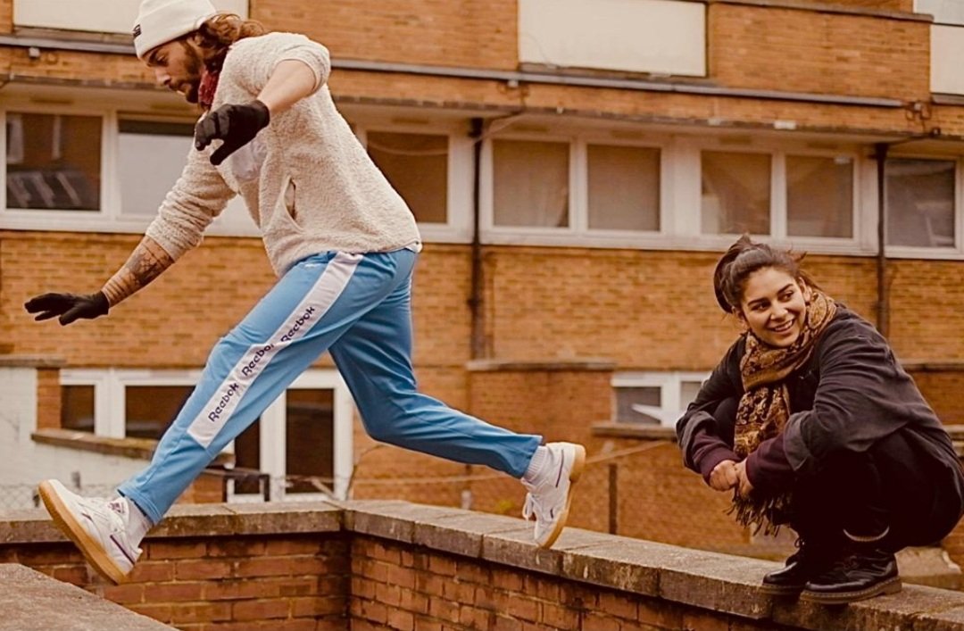 A young man in a beige sweater and blue pants is stepping off a brick ledge, with a woman crouching on the same ledge smiling. They are outdoors in front of an apartment building.