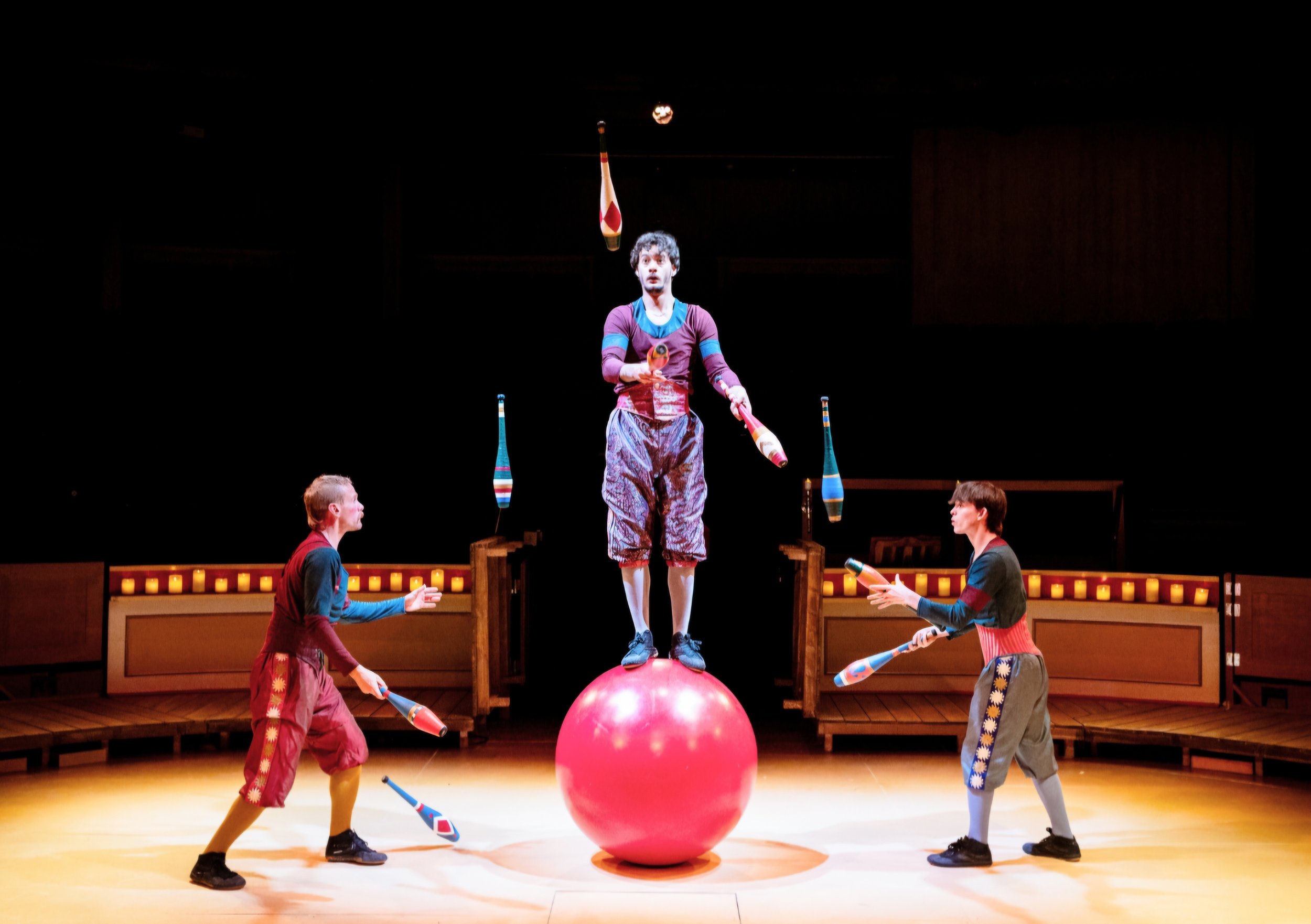 Circus performers juggling colorful clubs on stage, with one performer balancing on a large red ball in the center.