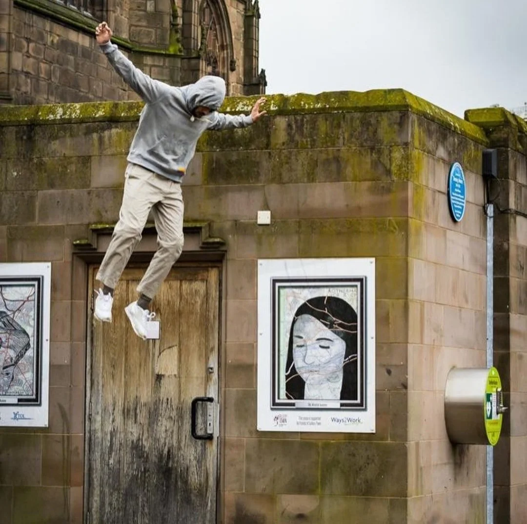 A person in a gray hoodie and beige pants appears to be falling or jumping off a moss-covered stone wall onto a weathered wooden door, in an urban setting with poster art and street signage visible.