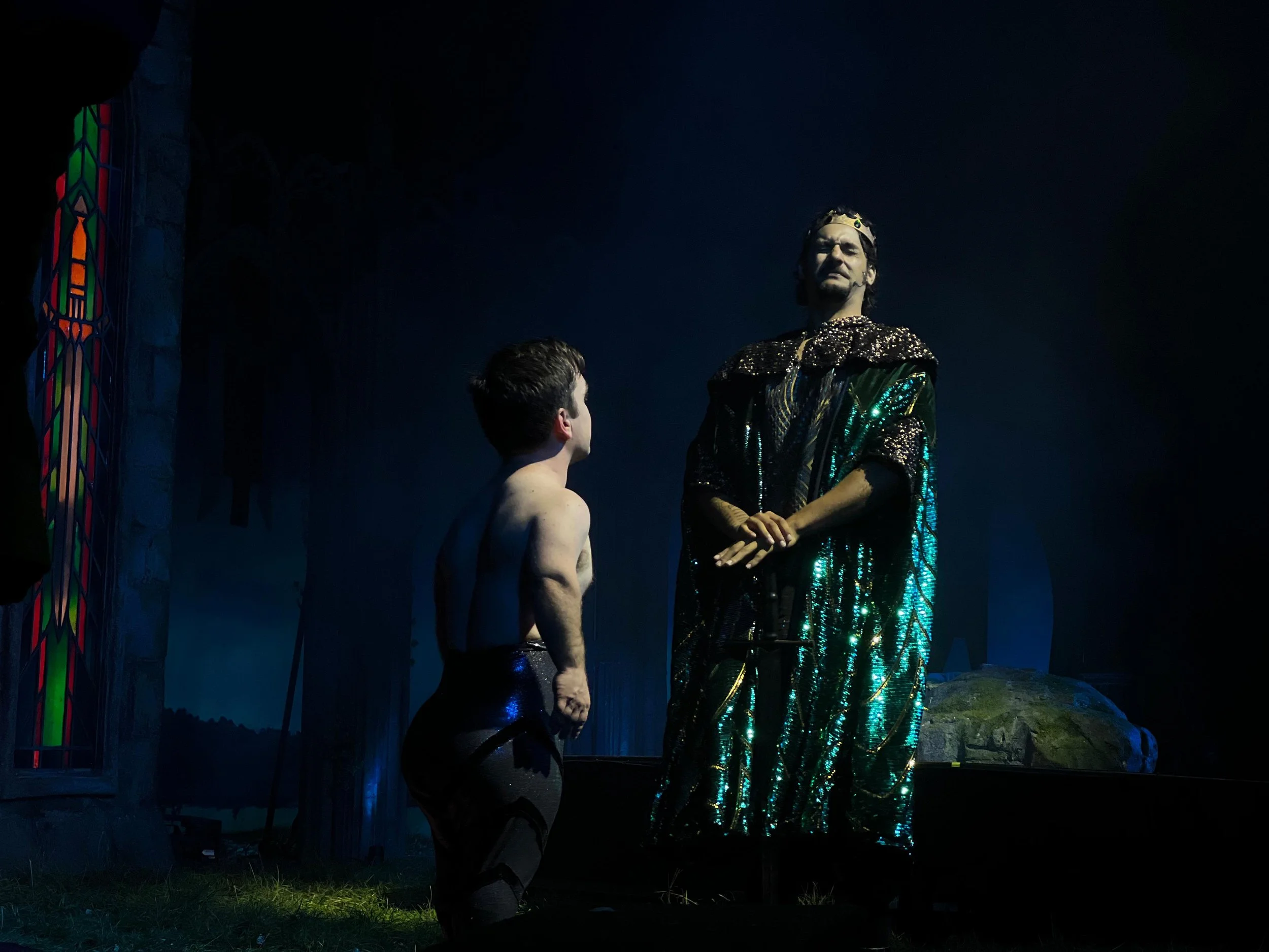 A scene from a theatrical performance featuring two characters: a young shirtless boy with dark pants looking up at an older man wearing a sparkling green and black robe with a crown, standing on a stage with dark background and colorful stained glas