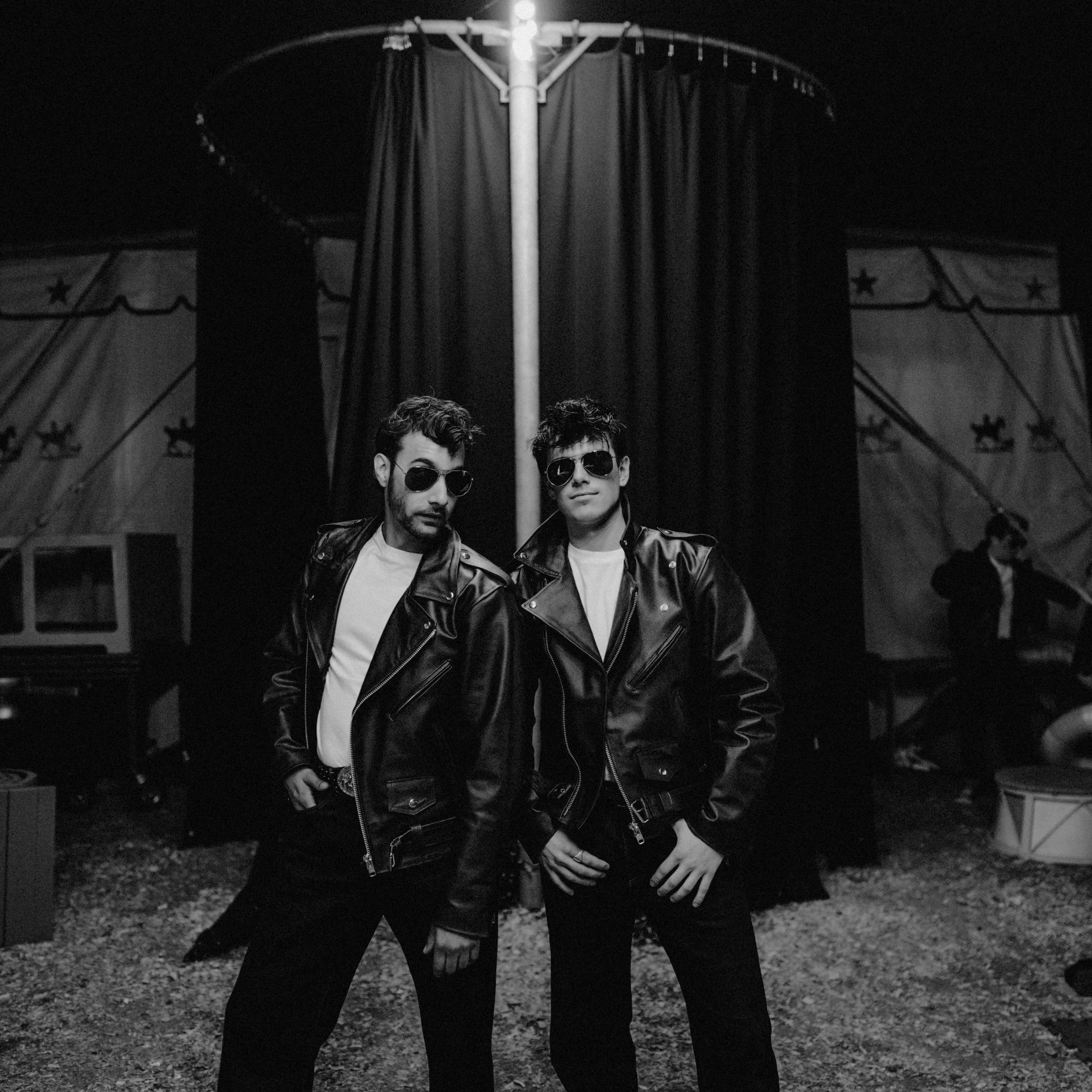 Two young men in leather jackets and sunglasses standing in front of a circus tent at night.