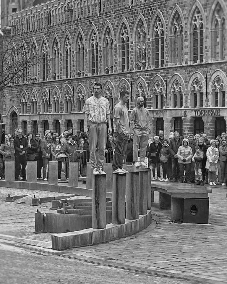 Three men stand on wooden posts in front of a crowd on a city street, with old brick-style buildings in the background.