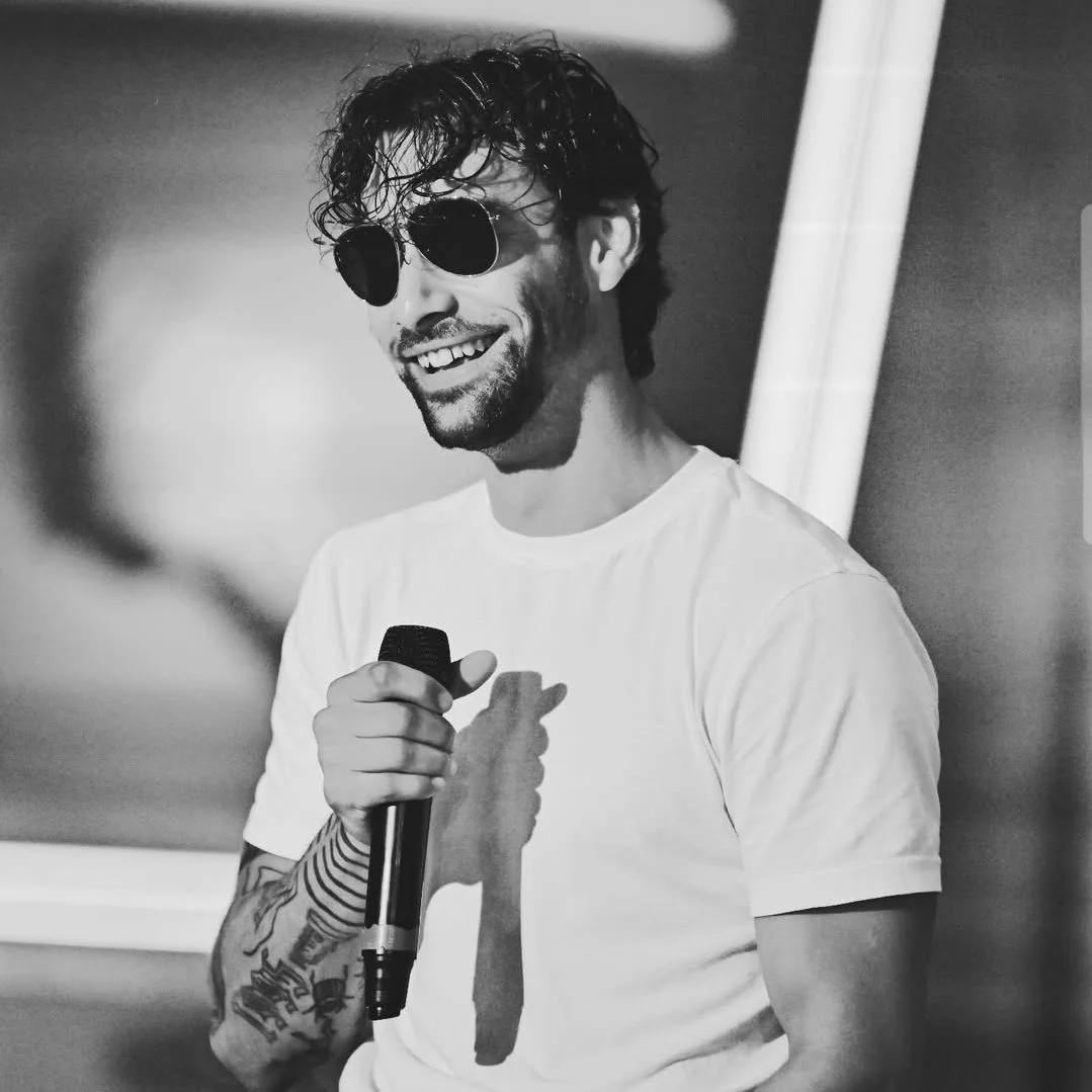 A man with curly hair, wearing sunglasses and a white t-shirt, smiling and holding a microphone.