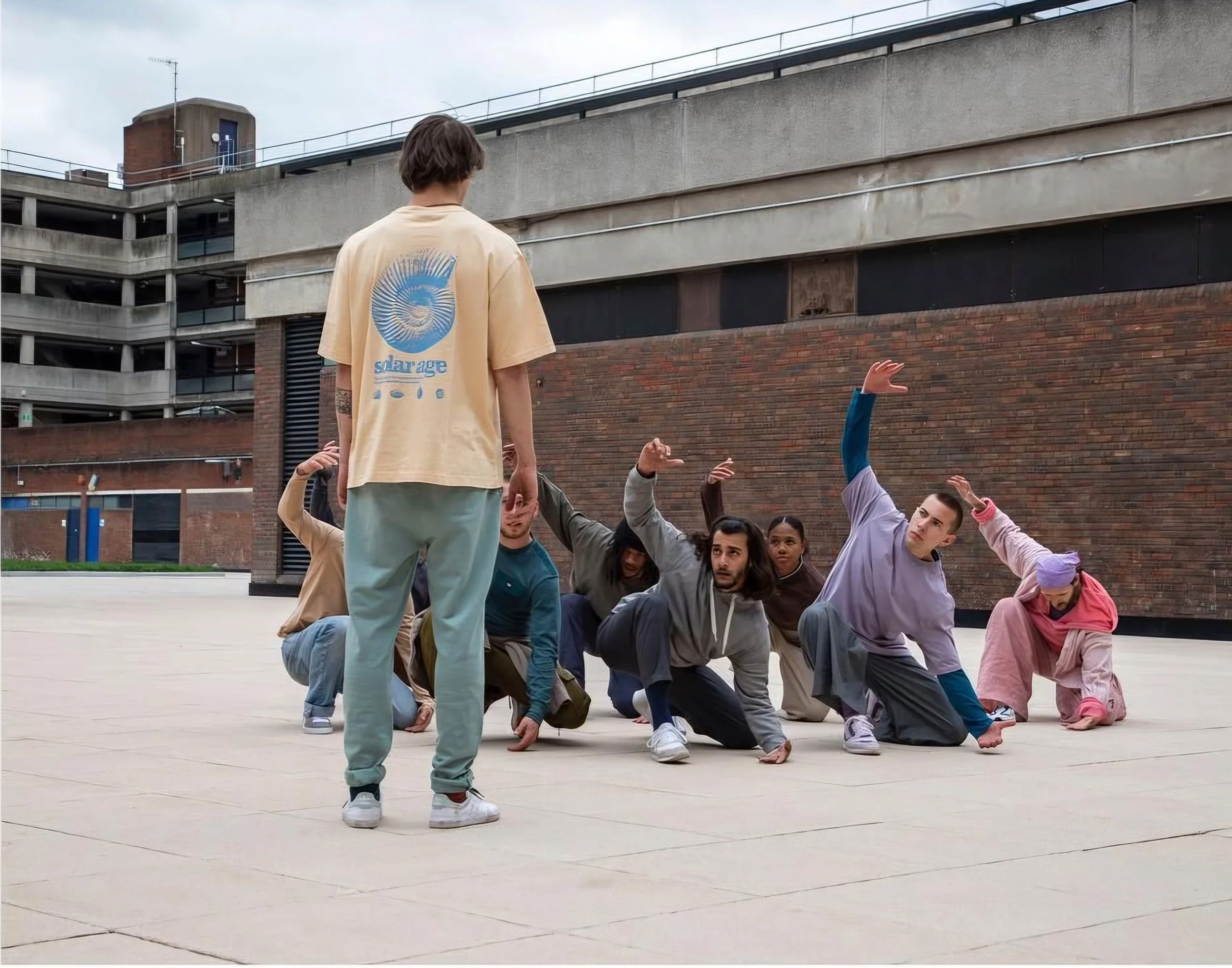 A group of seven diverse young people practicing breakdancing or hip-hop dance moves outdoors on a concrete plaza, with some crouching or leaning and raising their arms, while one person in front stands facing them with their back to the camera weari