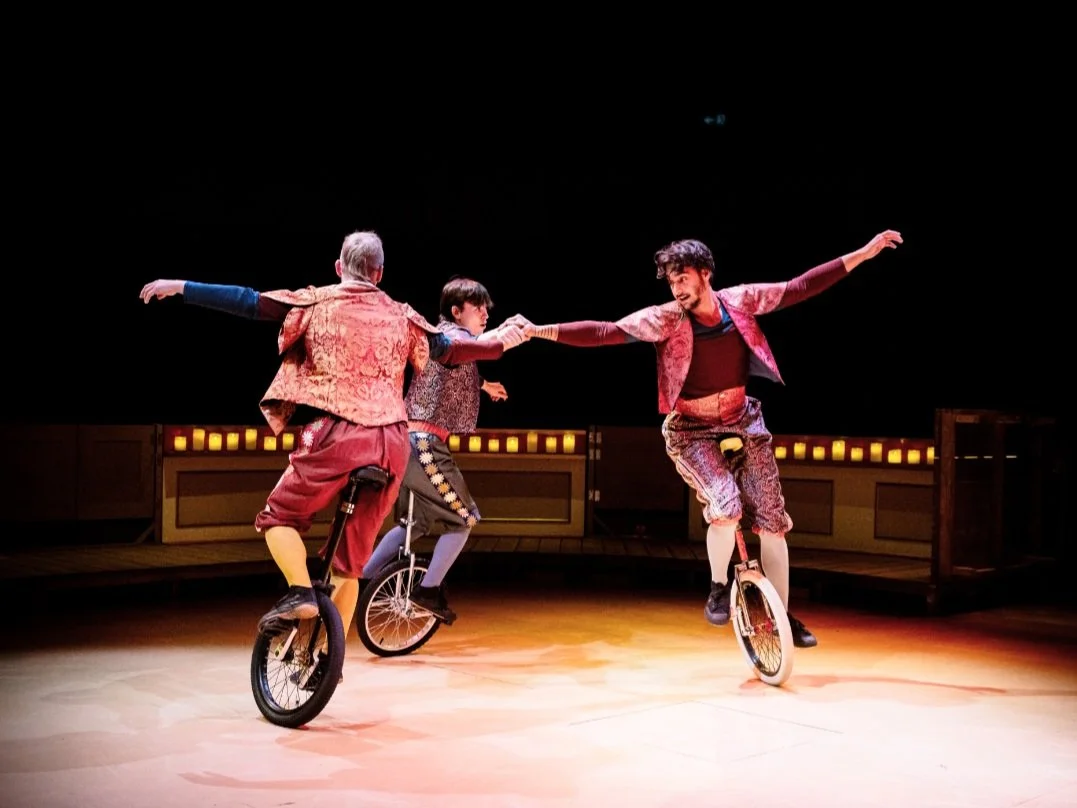 Three performers balancing on unicycles and holding hands during a stage act, wearing colorful costumes with a dark background and warm lighting.