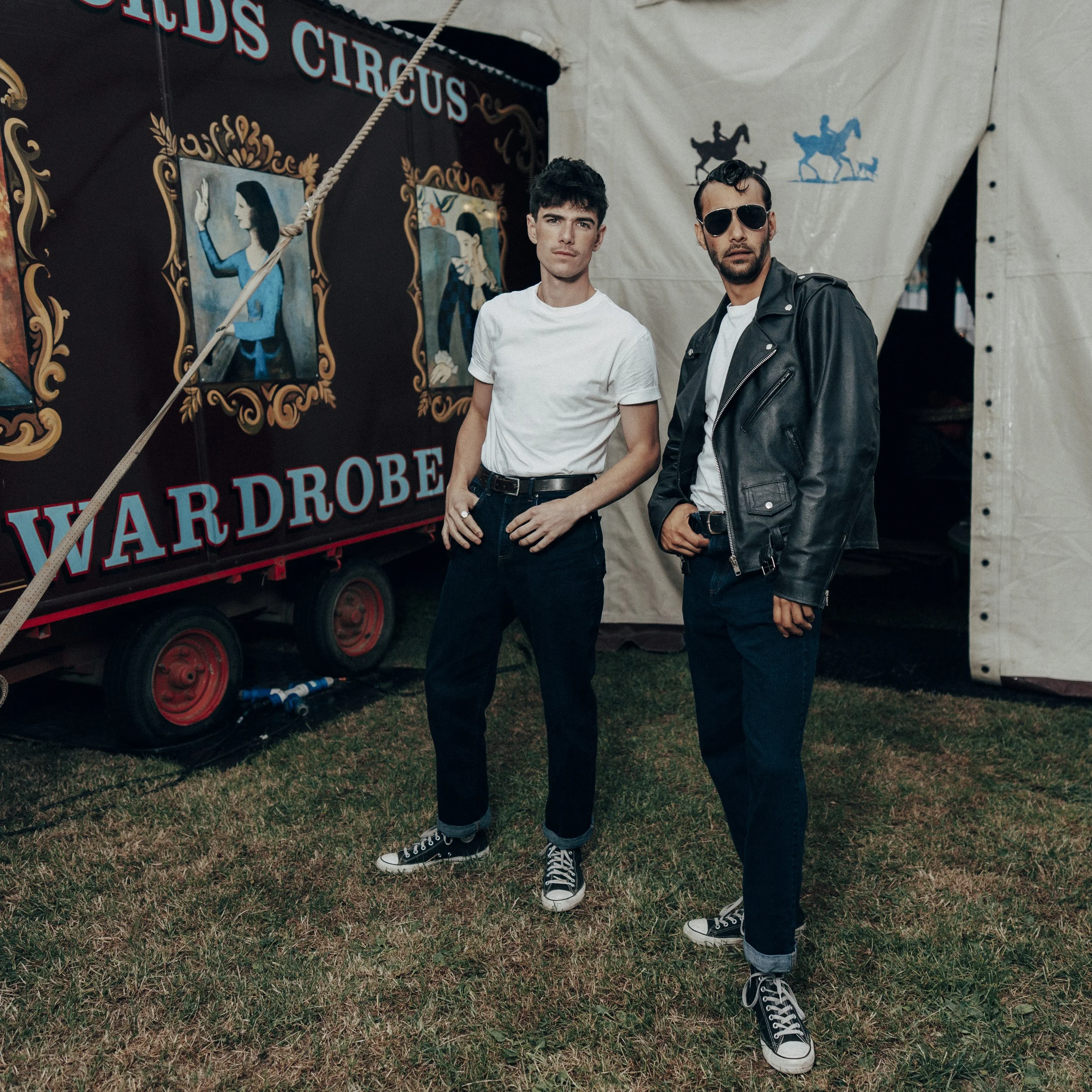 Two young men standing outside a circus tent, with a vintage-style poster behind them that reads "Freds Circus Wardrobe." One man wears a white t-shirt, black jeans, and sneakers, while the other wears a black leather jacket, white t-shirt, sunglasse