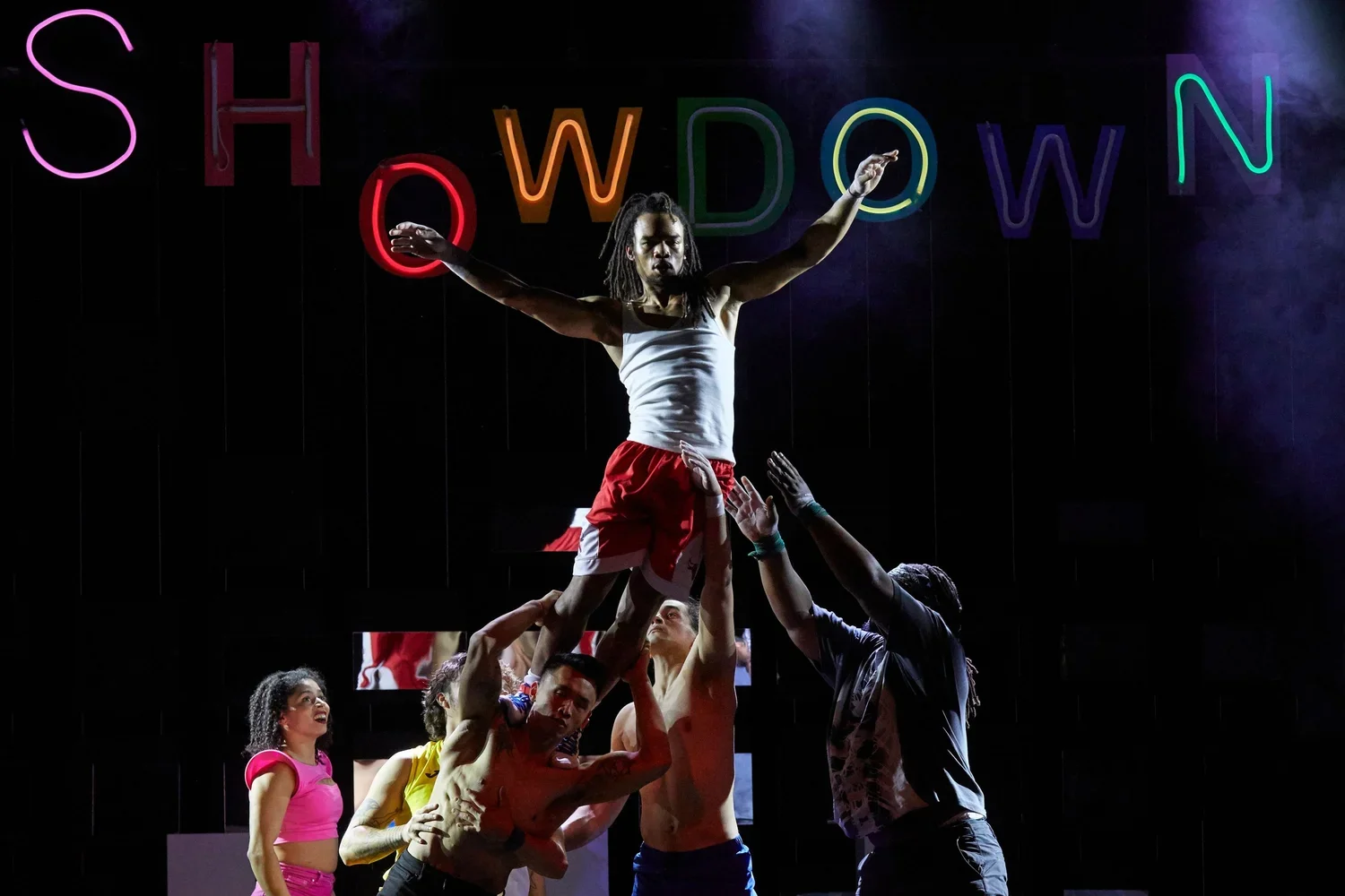 A group of people performing a human pyramid with one person standing on top, wearing a white tank top and red shorts, in front of a neon sign spelling 'SHOWDOWN'.