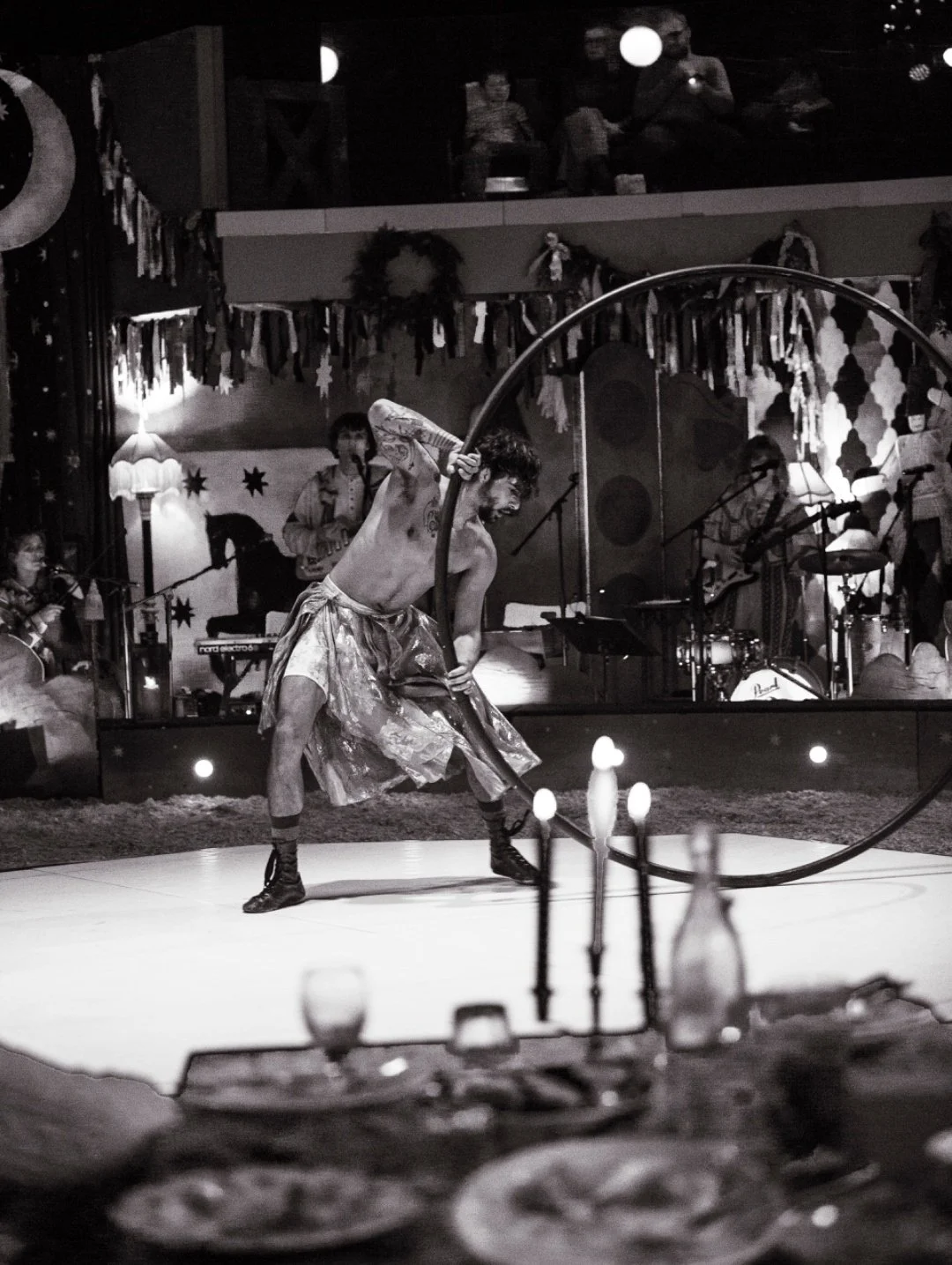 A shirtless performer with tattoos, wearing baggy pants and combat boots, performs an acrobatic move with a large hoop on a stage decorated for a festive event, with musicians playing in the background.