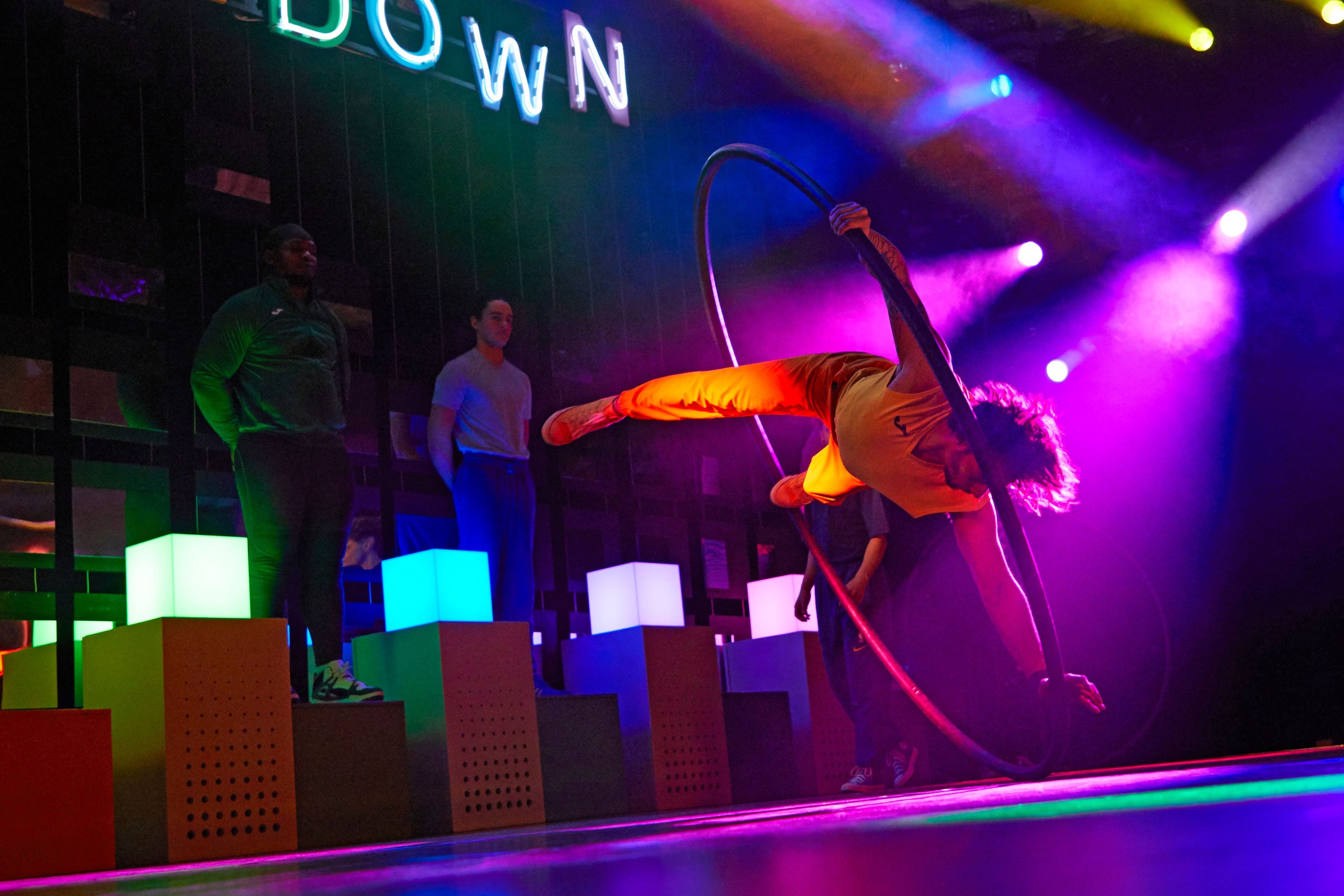 A circus performer with curly hair is balancing on a large ring while holding another ring, in front of a colorful, illuminated background with three other people watching.