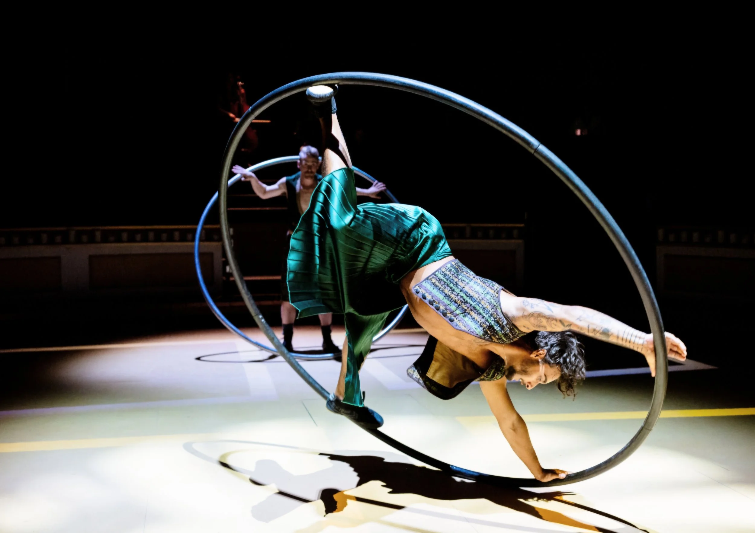 A performer in a green costume balances on a Cyr wheel during a circus act, with another performer in a black dress in the background.
