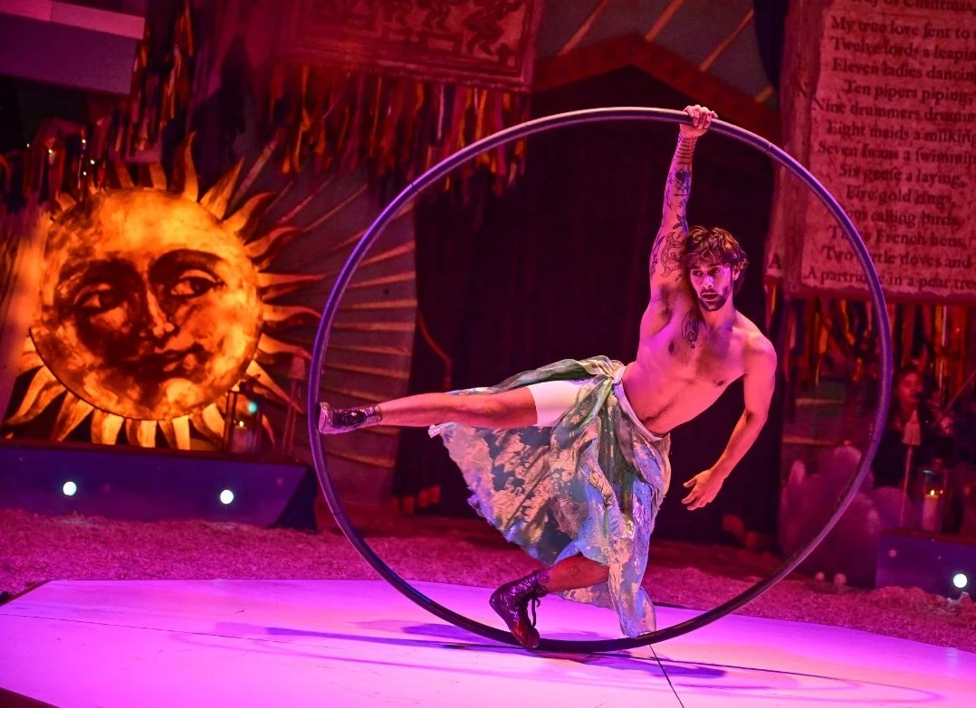 A shirtless male circus performer with tattoos on his arm and chest performs on a Cyr wheel, standing on one foot inside the wheel, with a colorful circus backdrop featuring a sun with a face and what appears to be a poem banner in the background.