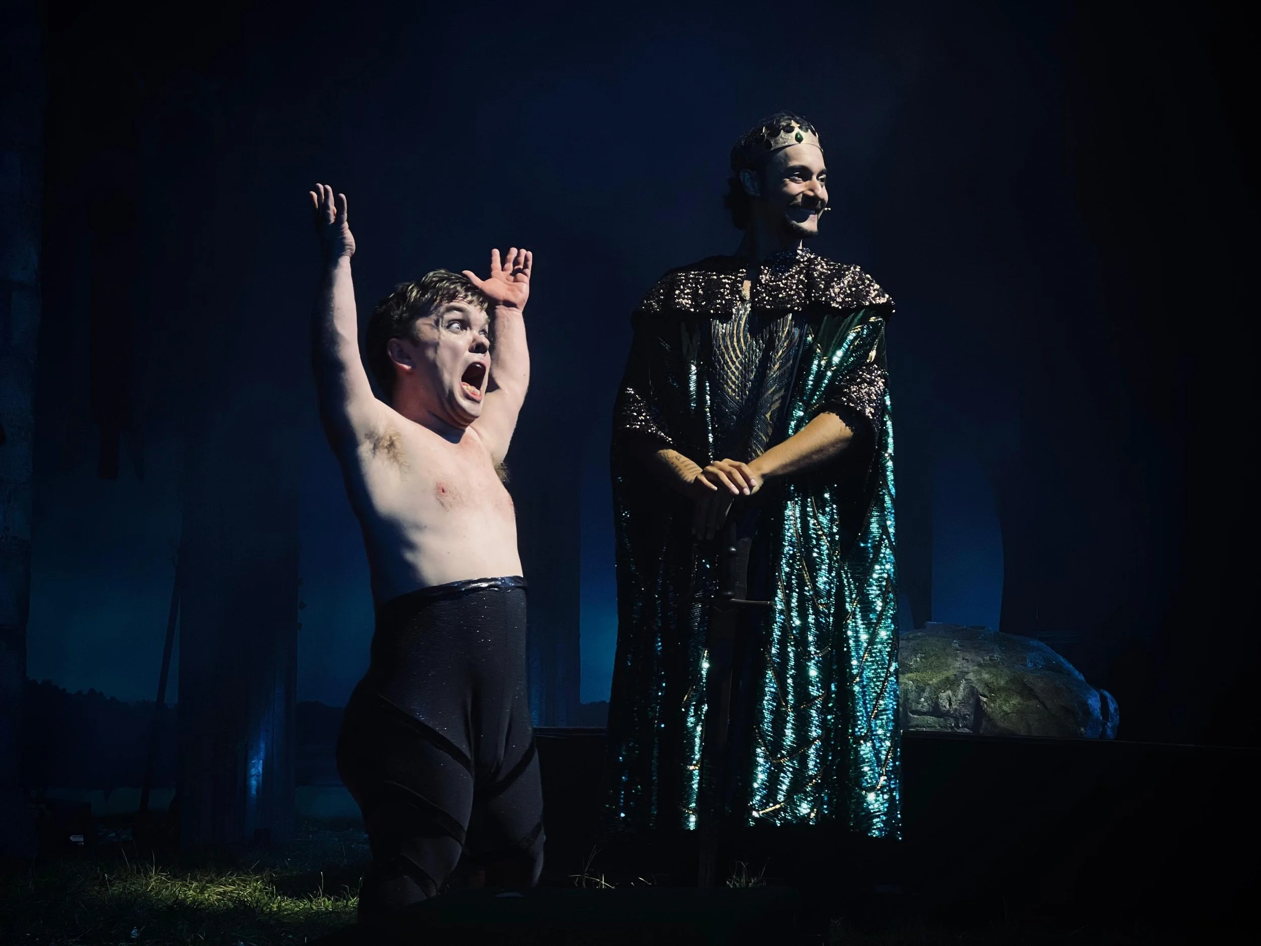 Two male actors on stage in theatrical costumes, one shirtless with a distressed expression and the other wearing a shiny, ornate robe and crown, in a dark forest setting.