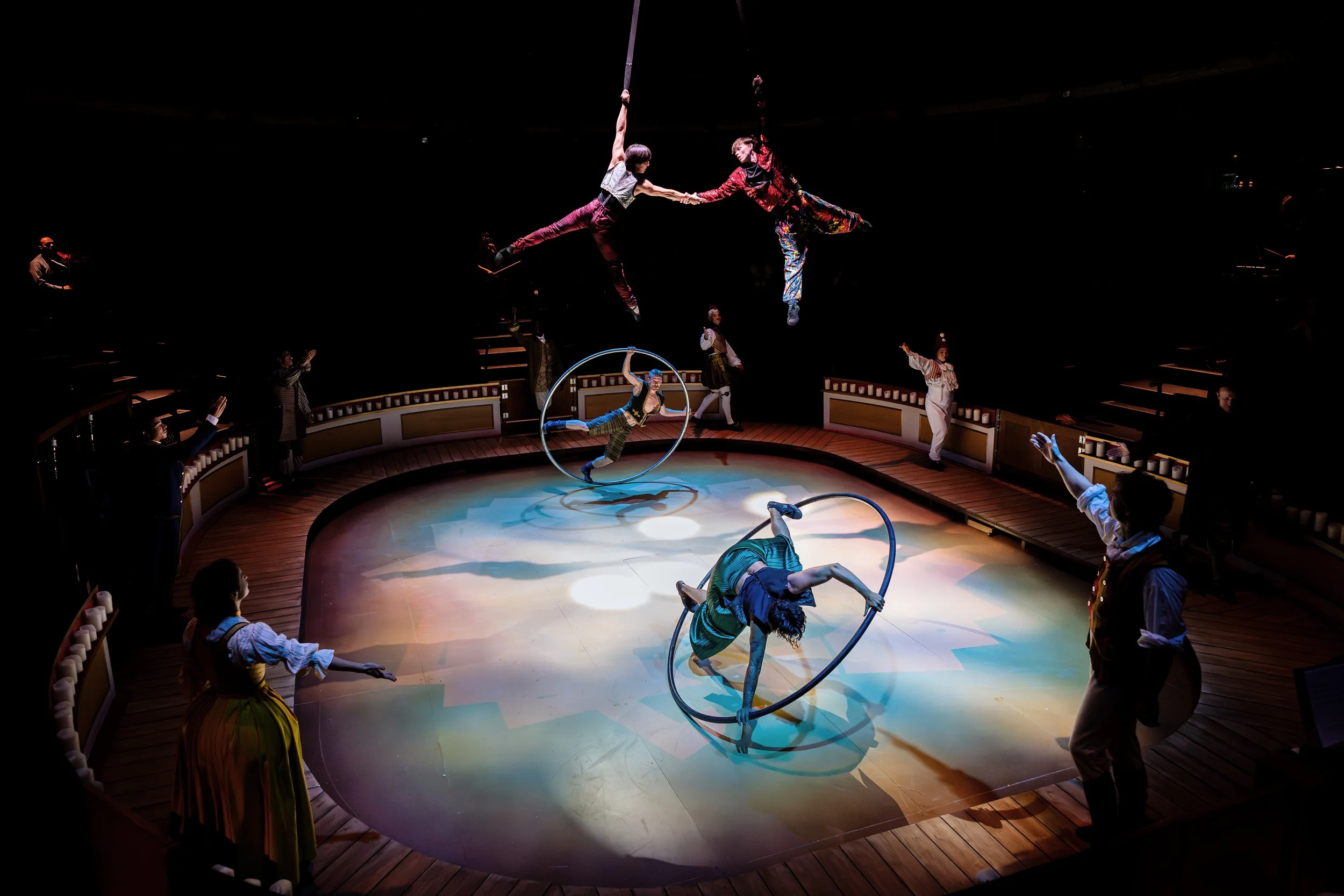 Circus performers demonstrating acrobatics and aerial acts on a circular stage, with audience members and assistants surrounding the stage.