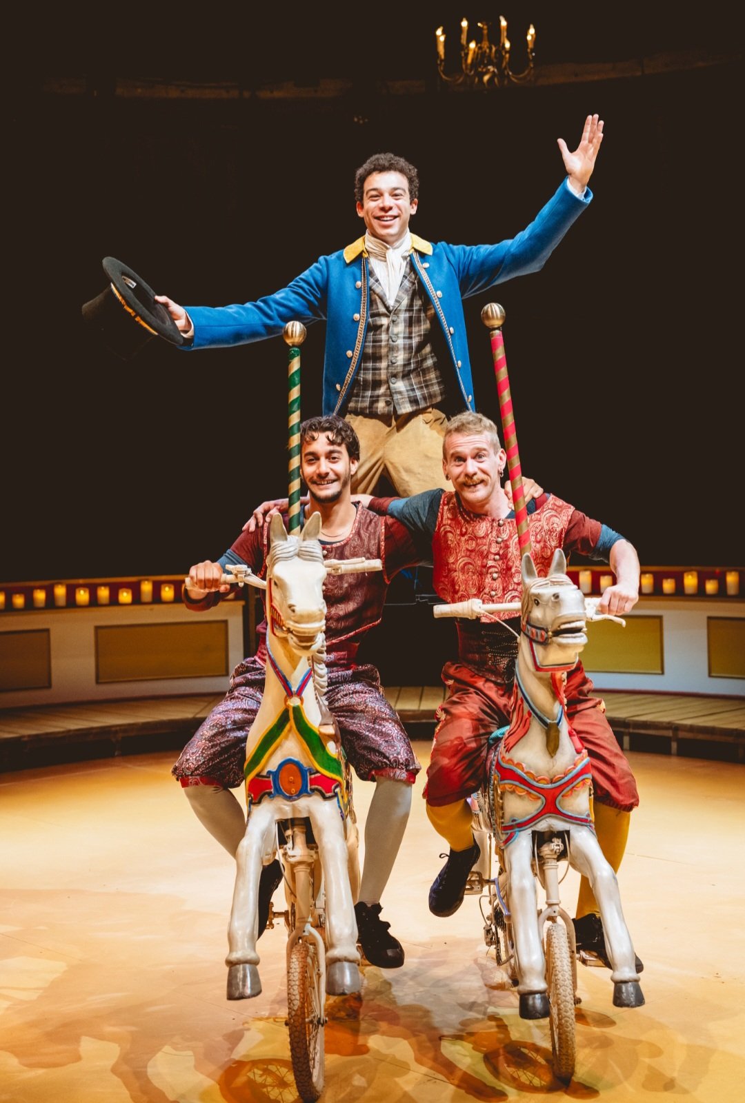 Three male performers in colorful costumes riding toy horses on a circus stage, with one performer standing on the shoulders of the other two. The performer at the top is smiling and holding a hat, while the two others are riding the horses and smili