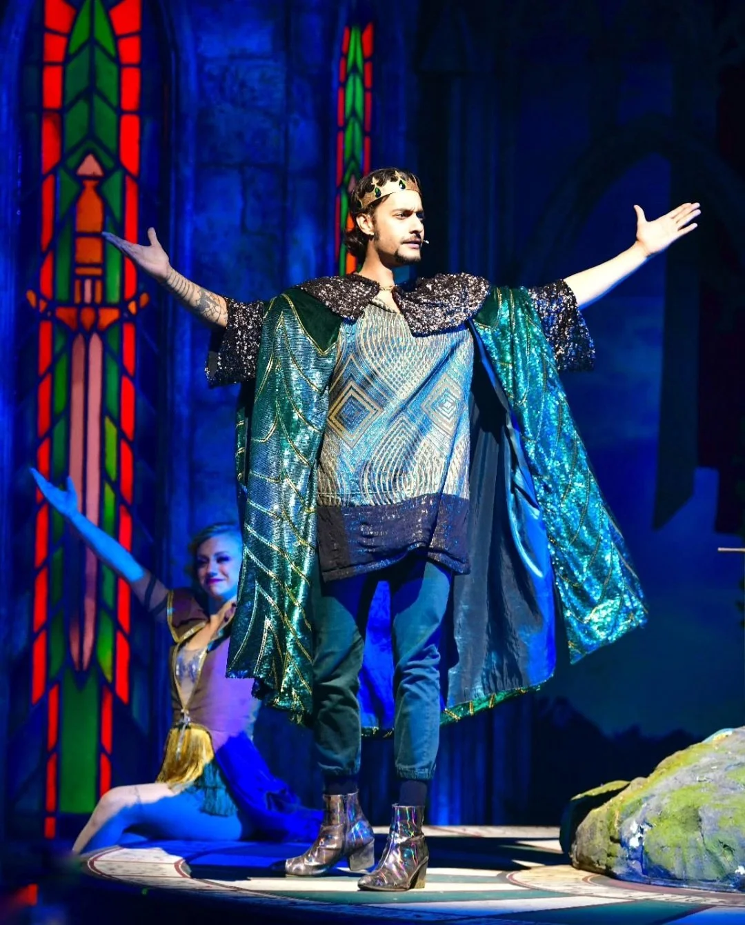 A stage performance featuring a male actor dressed as a king or wizard with arms outstretched, wearing a crown, ornate cape, and intricate clothing. A female performer is in the background, sitting on the stage with one arm raised. The set includes c