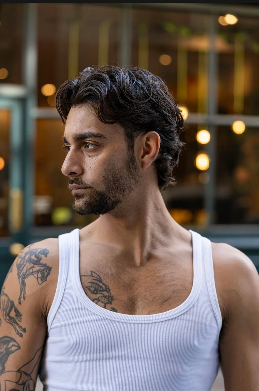 A man with dark wavy hair and a beard wearing a white tank top, showing tattoos on his shoulder and chest, standing outdoors with blurred warm lights in the background.