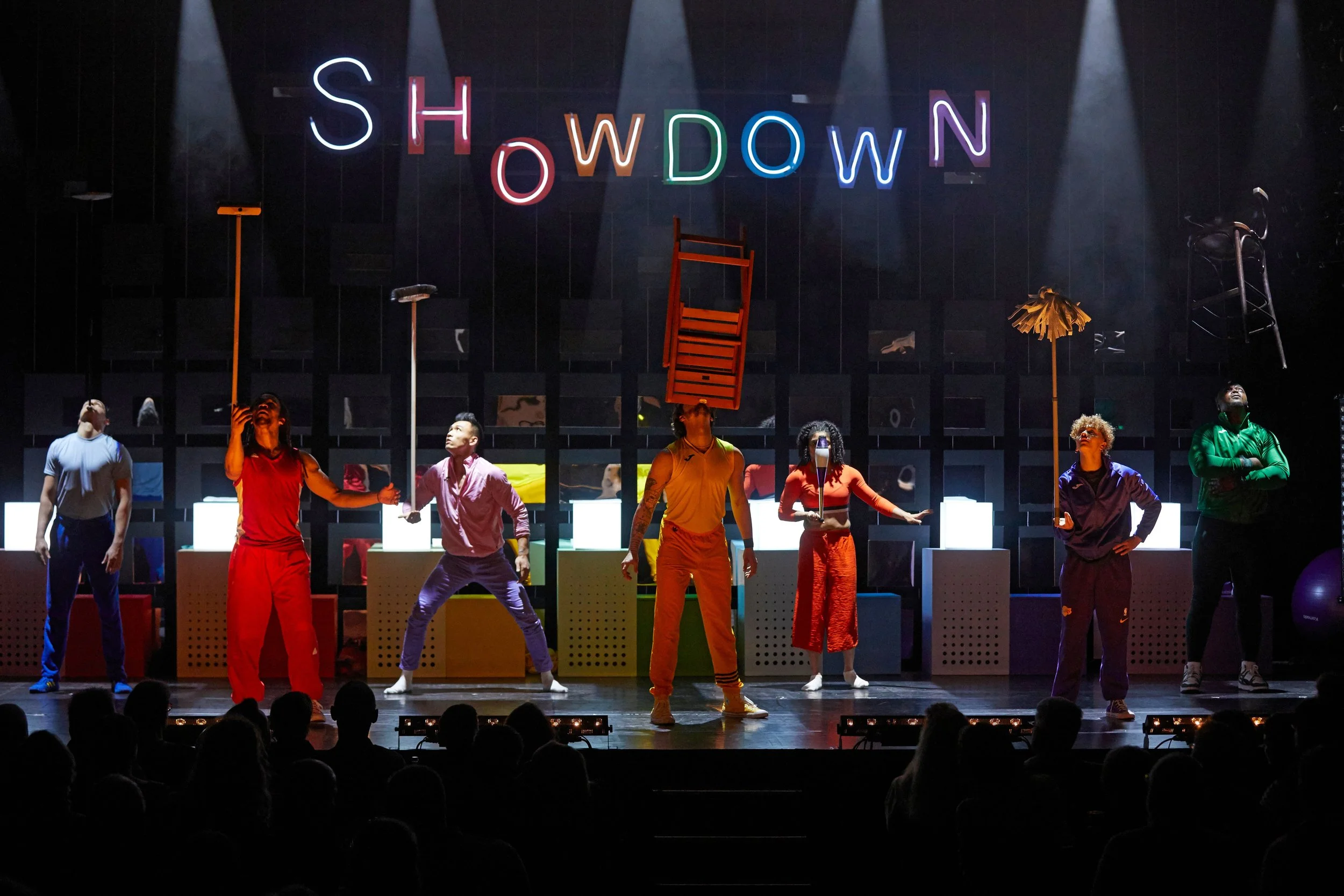 A stage performance with six performers rehearsing or performing a show with props such as chairs, umbrellas, and other objects hanging or balanced. The background features the word 'SHOWDOWN' spelled out in colorful, illuminated letters. Audience me