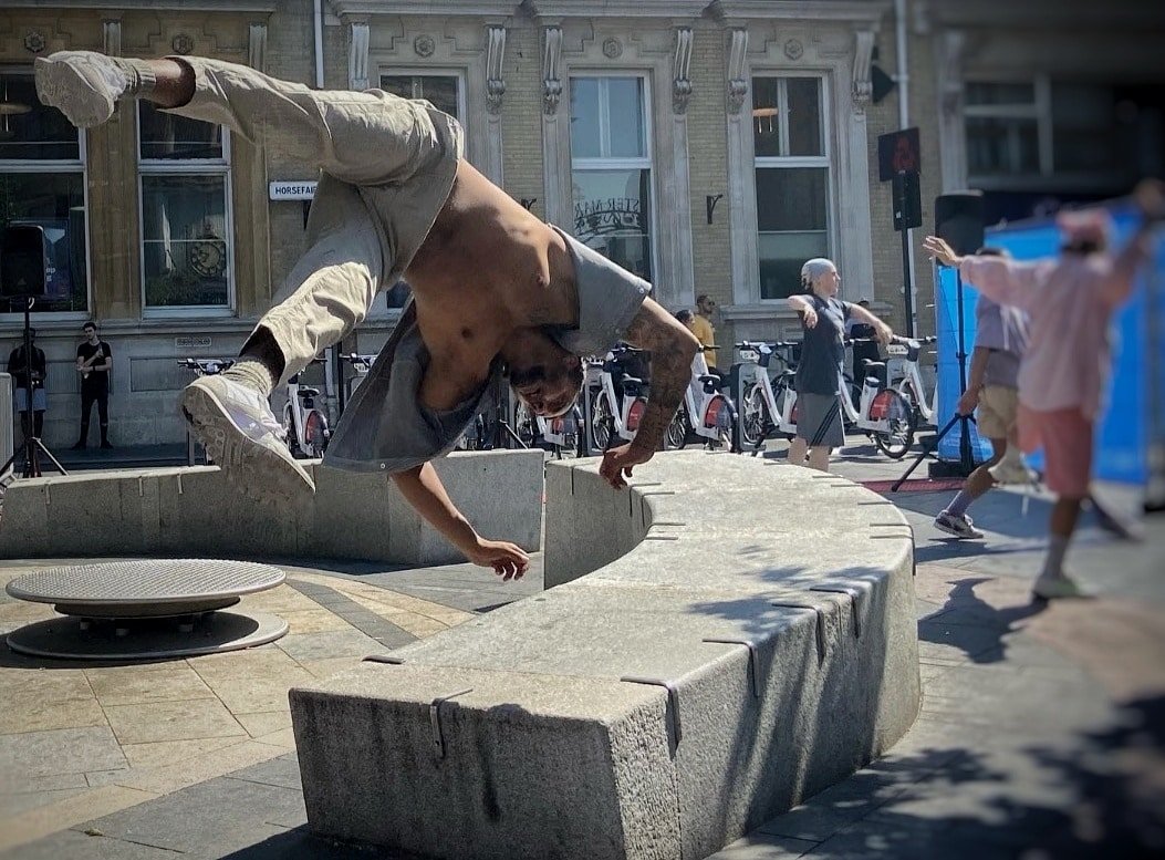 A person performing a breakdancing move on a rounded stone bench in an urban setting with pedestrians and bicycles in the background.