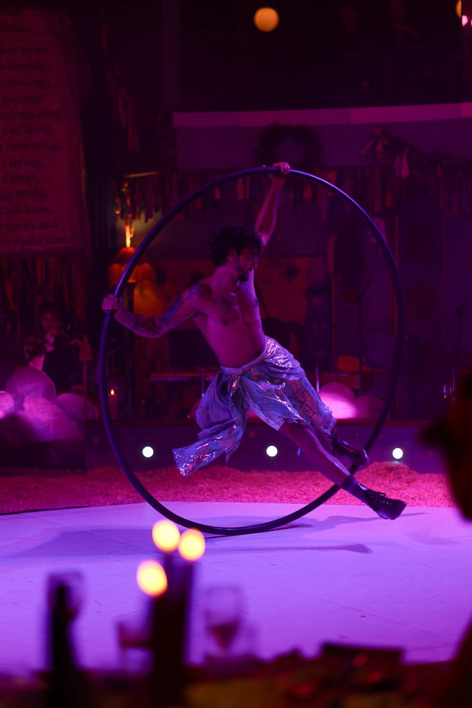 A shirtless male acrobat performing with a Cyr wheel in a circus setting with dim purple and pink lighting.