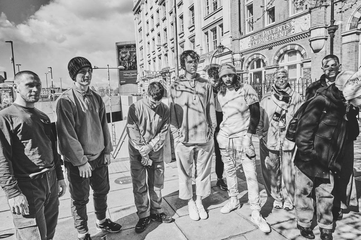 Group of people standing outside in an urban environment, some wearing hoodies and jackets, with a large building in the background. It's a black-and-white photo showing clear weather and shadows from trees.
