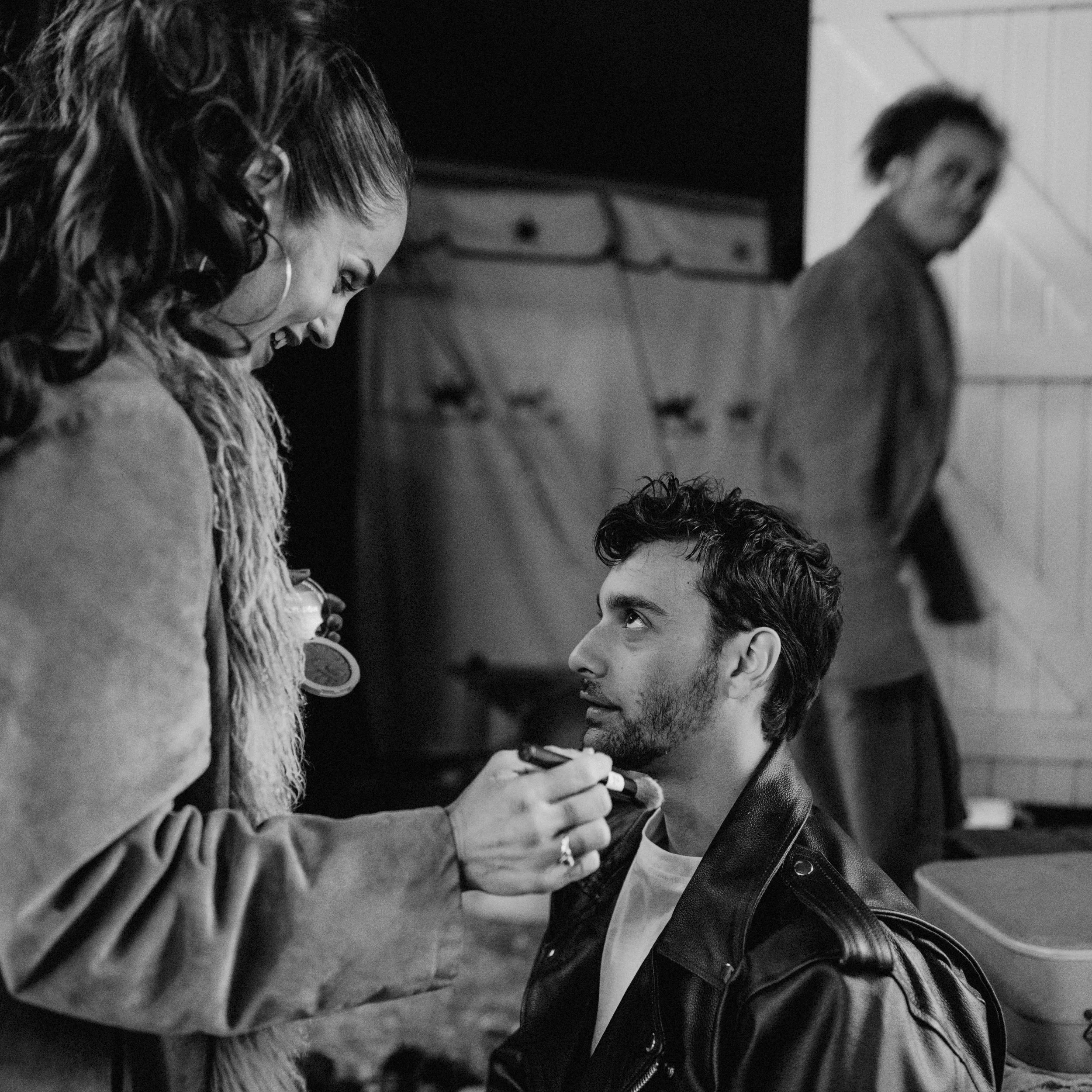 A woman is applying makeup to a man seated in front of her, with an expressive look on her face, indoors with another person in the background.