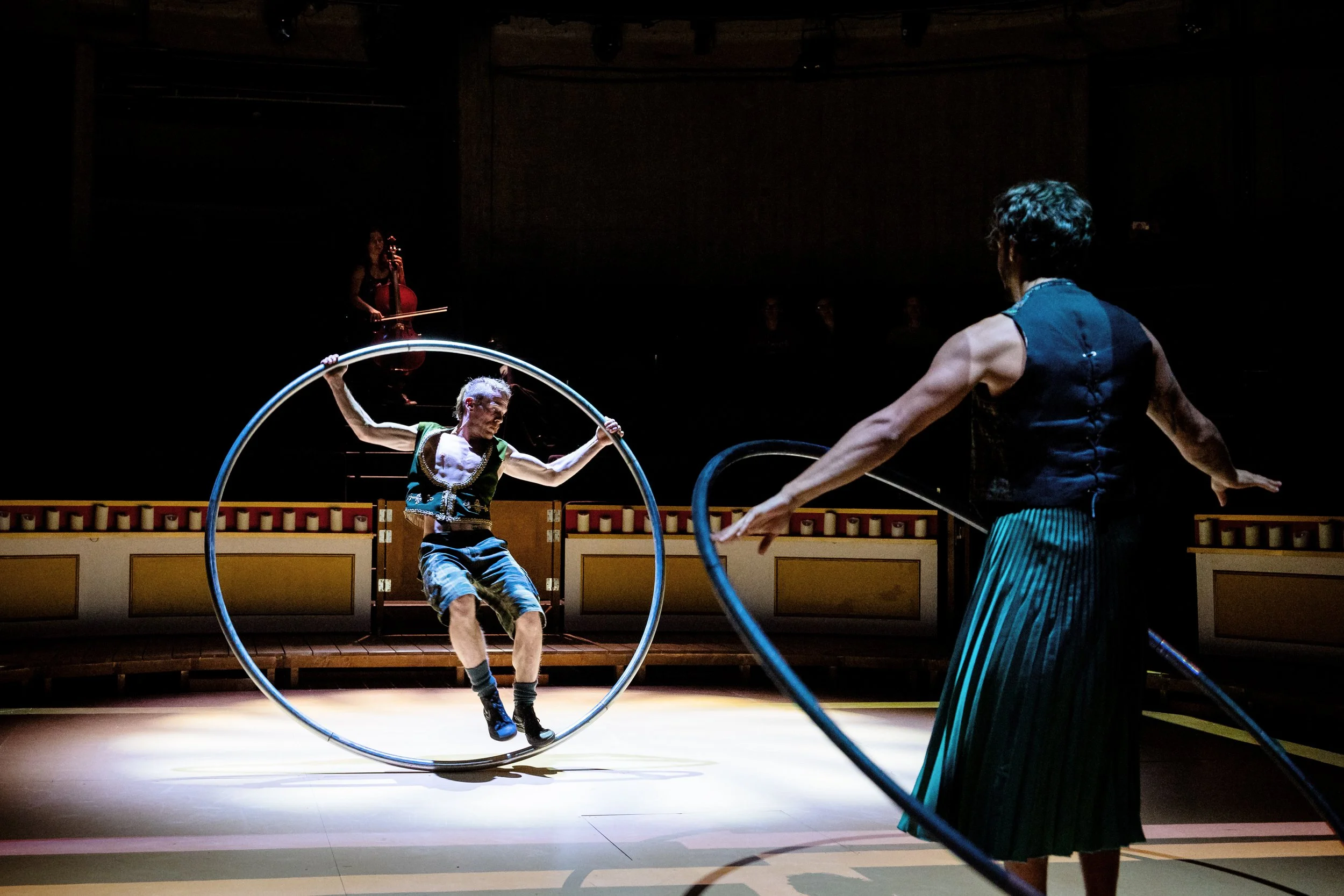Two performers in a circus show, one spinning inside a metal hoop and the other holding a hoop, in a dimly lit indoor venue with a musician playing violin in the background.