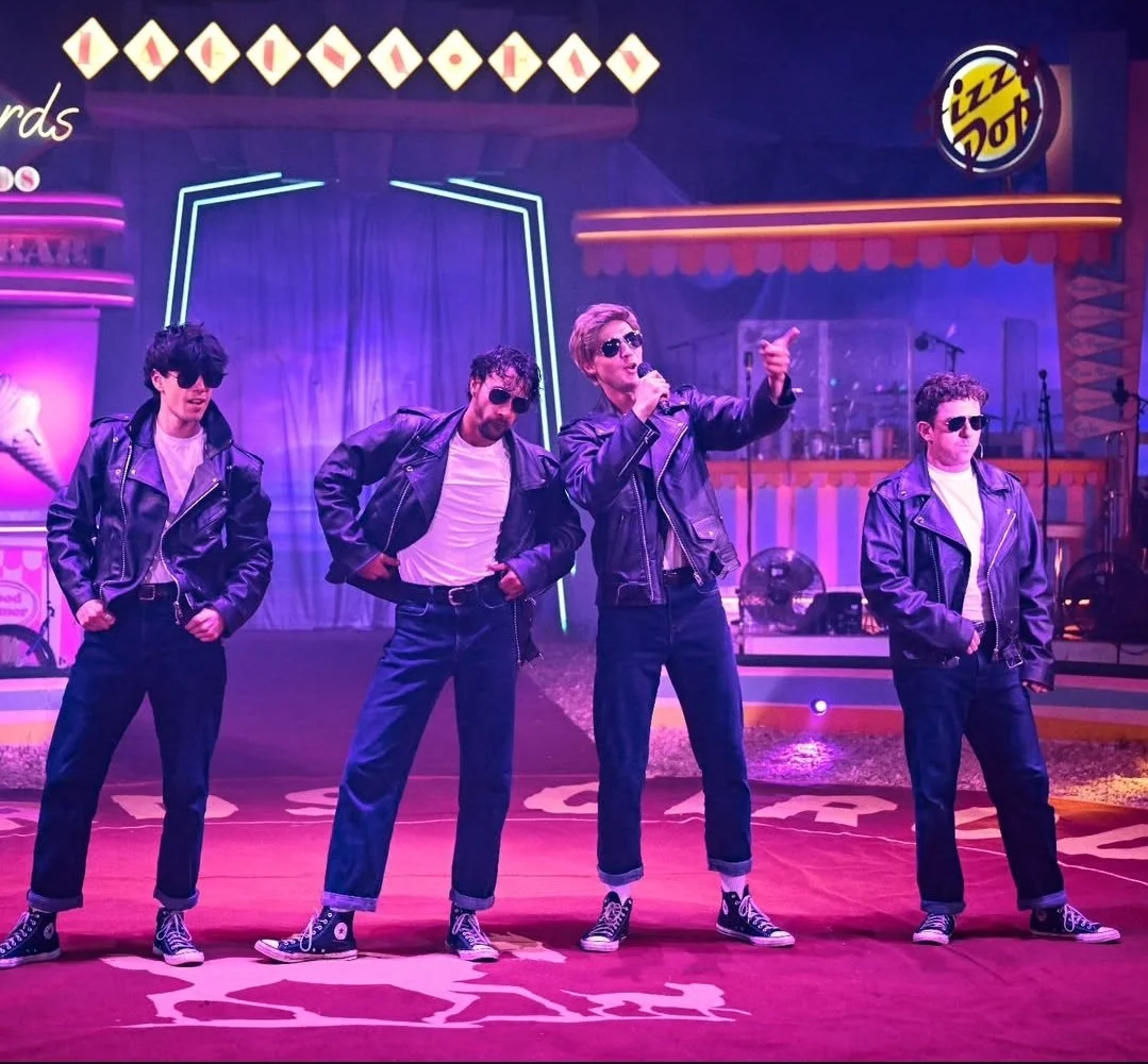 Four young men dressed in black leather jackets, sunglasses, white T-shirts, and black jeans performing on stage with a 1950s diner-themed background.