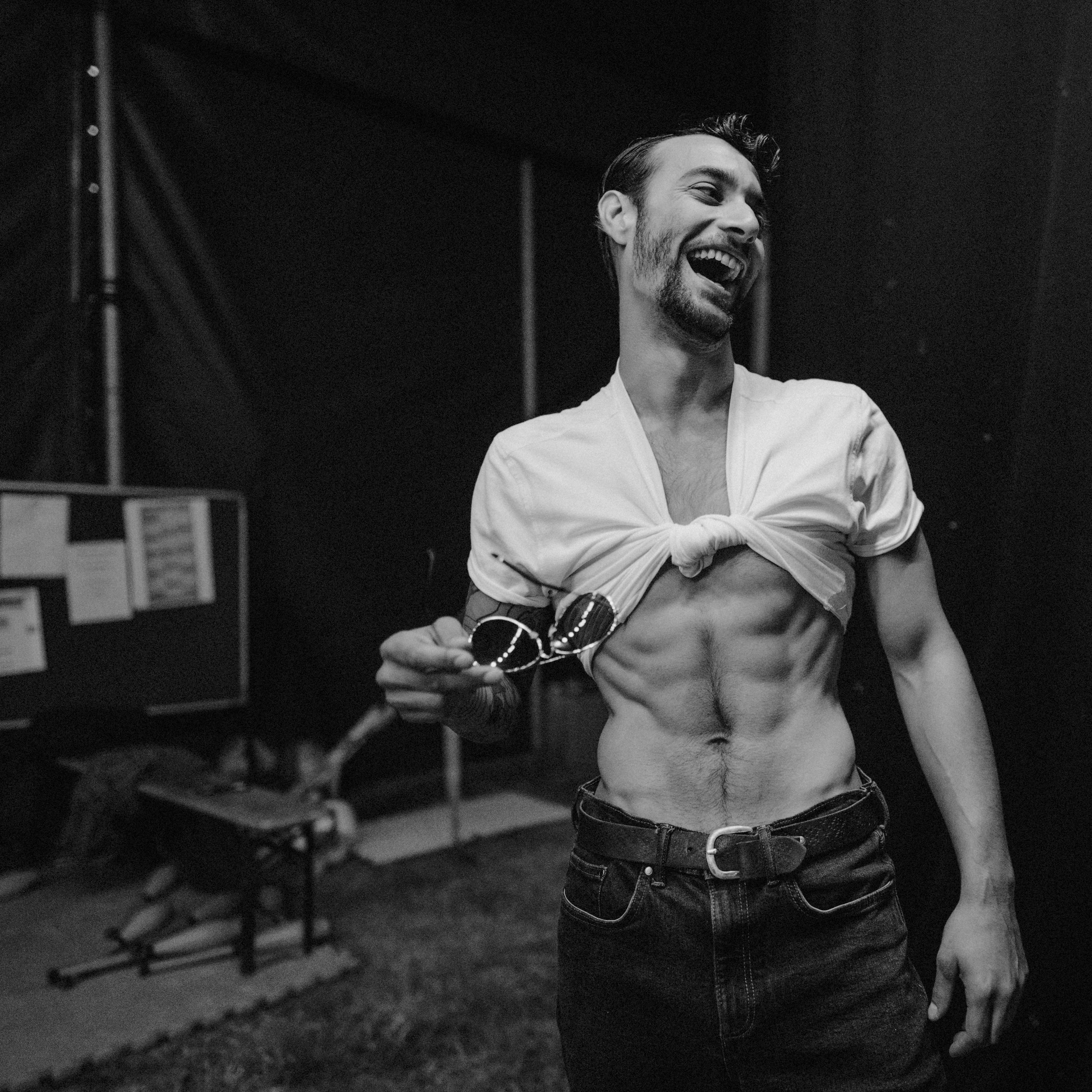 A man with a beard and styled hair smiling, lifting his tied-up shirt to reveal his abs, holding sunglasses, in a dimly lit room.