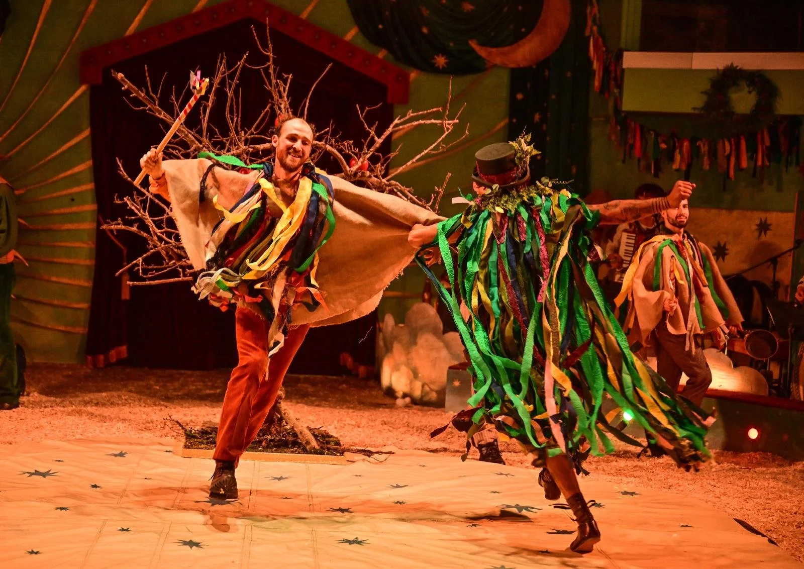 Two performers in colorful costumes dancing on stage with a nature-themed background. One dancer wears a cape made of branches and colorful ribbons, and the other wears a dress with green and yellow ribbons, and a hat. Small trees and festive decorat