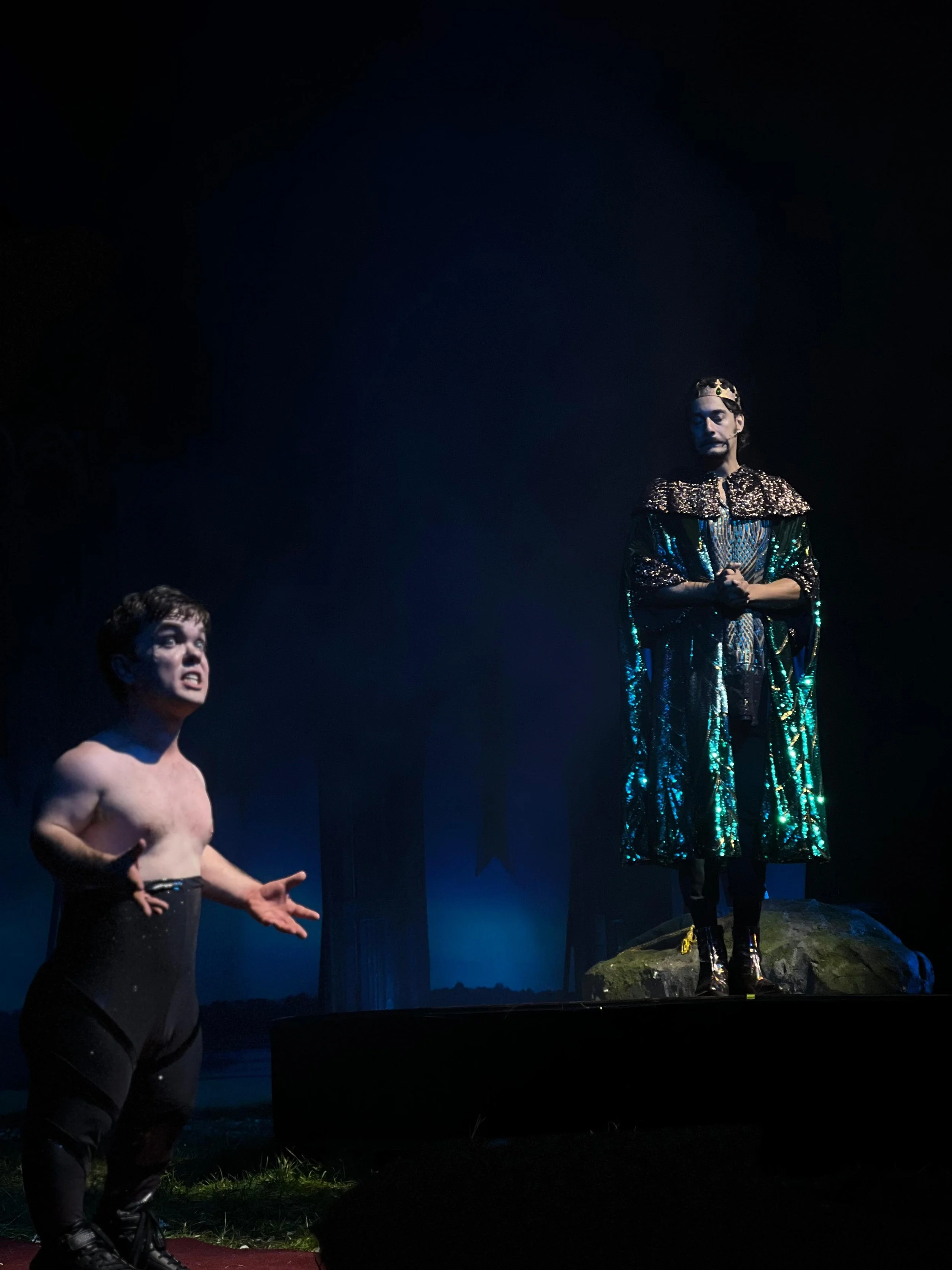 A scene from a theater performance with two actors on stage. One actor, a shirtless man wearing black pants, appears to be speaking or expressing emotion. The other actor, dressed in a shiny cape and a crown, stands on a rock, looking contemplative.