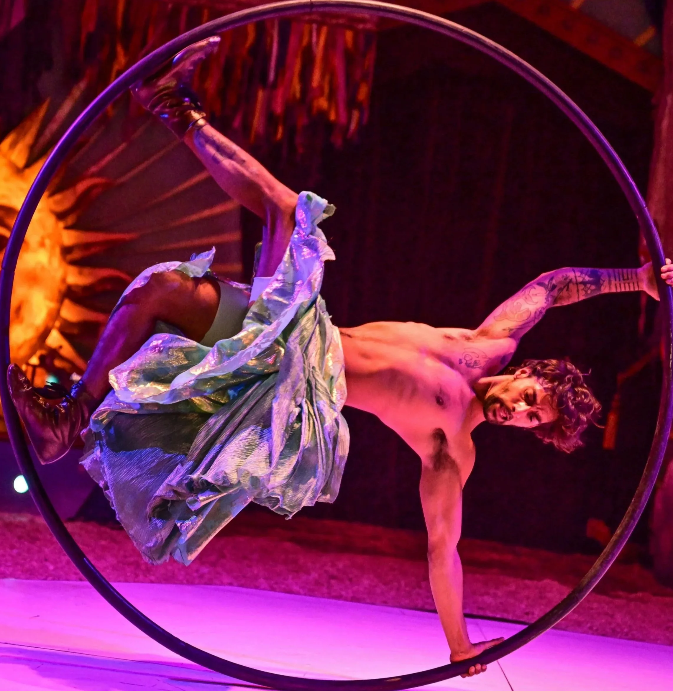 A shirtless man performing on a Cyr wheel, wearing shorts and shoes, inside a circus tent with orange and purple lighting.