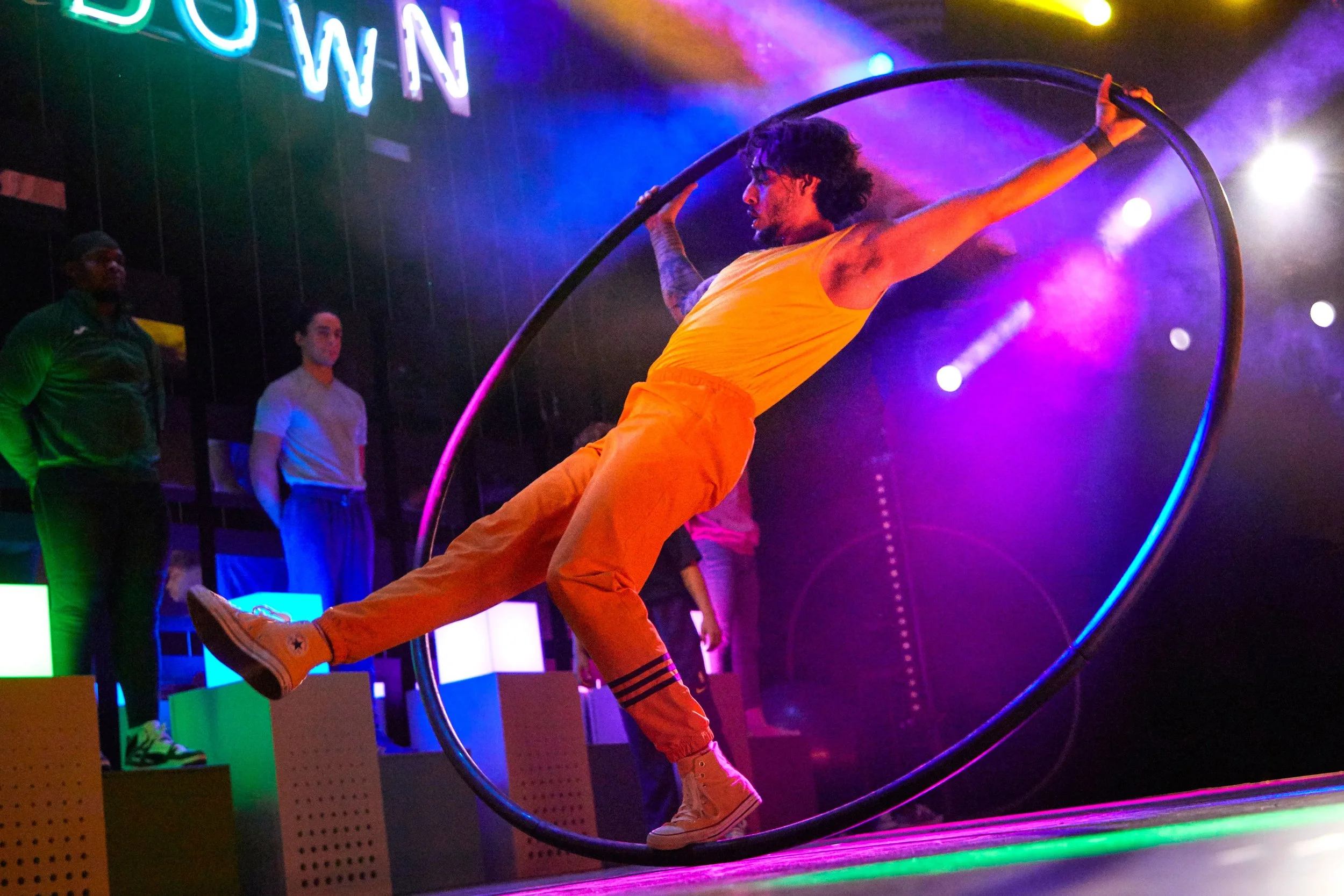 A performer in an orange outfit does acrobatics on a Cyr wheel in a nightclub or entertainment venue with colorful neon lights and spectators in the background.