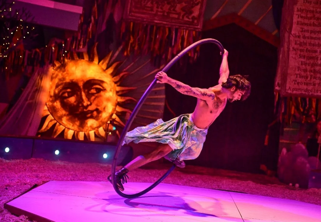 A male performer in a colorful skirt spins inside a metal ring on a circus stage. Behind him is a large sun with a smiling face painted on it, and the stage is decorated with vibrant banners and flags.